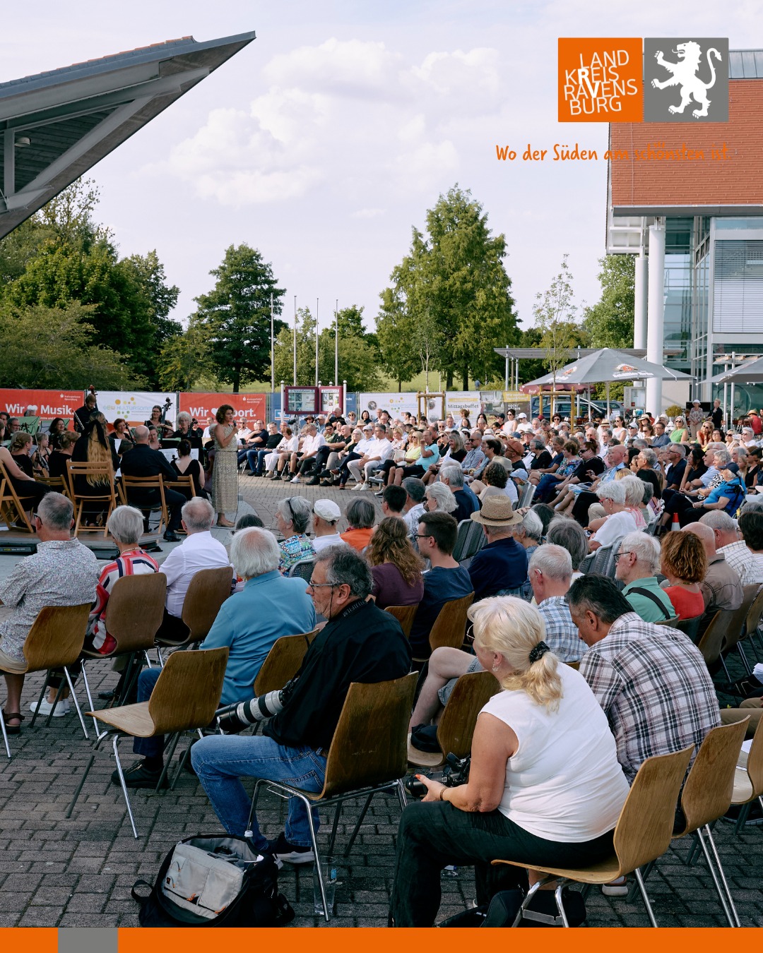 Eine große Gruppe von Menschen sitzt in Stühlen im Freien und schaut einer Person und einer Band auf der Bühne zu.