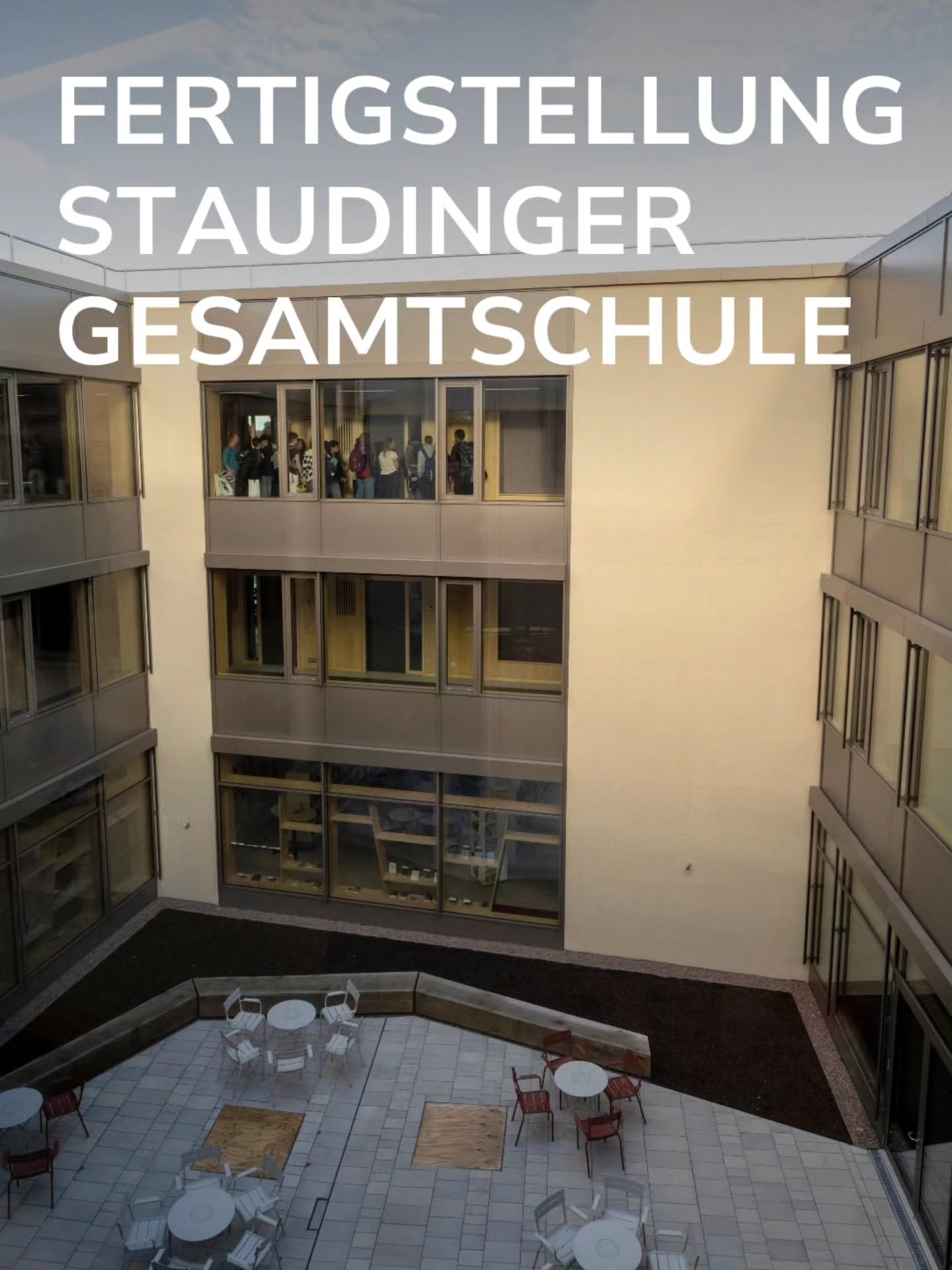 Ein gepflasterter Schulhof mit Tischen und Stühlen wird von einem modernen Gebäude mit großen Fensterfronten umgeben, in denen mehrere Personen zu sehen sind. Über der Szene prangt in großen weißen Lettern der Text „FERTIGSTELLUNG STAUDINGER GESAMTSCHULE“. (KI-Alternativtext)
