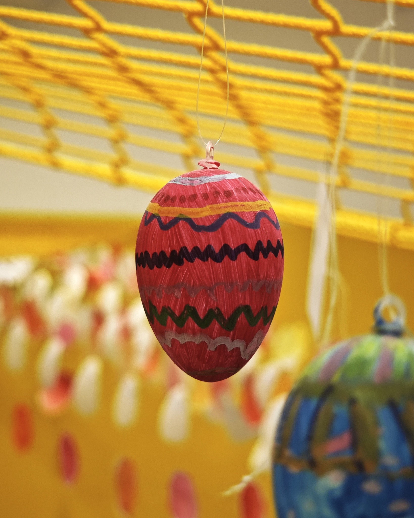 Dieser Alternativtext wurde KI-generiert: Ein roter, bemalter Gegenstand hängt von einer Schnur herab. Im Hintergrund ist eine gelbe Struktur und weitere dekorative Elemente zu erkennen.