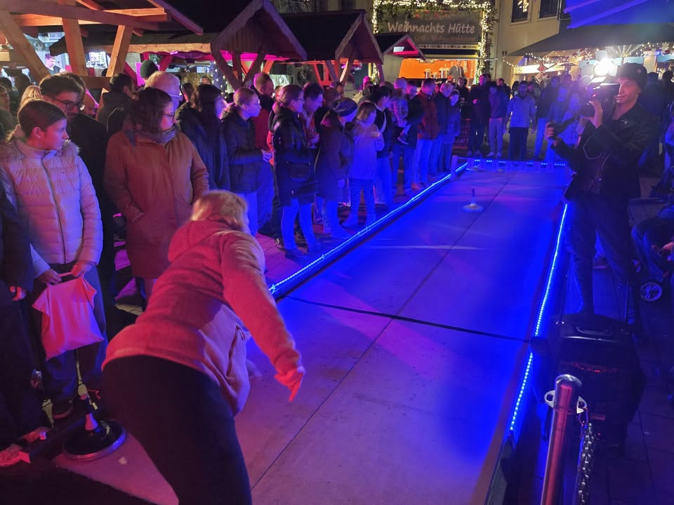 Dieser Alternativtext wurde KI-generiert: Eine Menschenmenge steht um eine beleuchtete Eisbahn herum. Im Hintergrund sind festlich geschmückte Buden zu sehen.