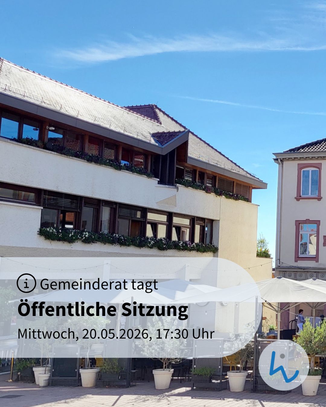 Gemeinderat tagt, öffentliche Sitzung am Mittwoch, 20.05.2026, 17:30 Uhr. Im Hintergrund ein mehrstöckiges Gebäude mit Blumenkästen und Terrasse unter Sonnenschirmen. (KI-Alternativtext)