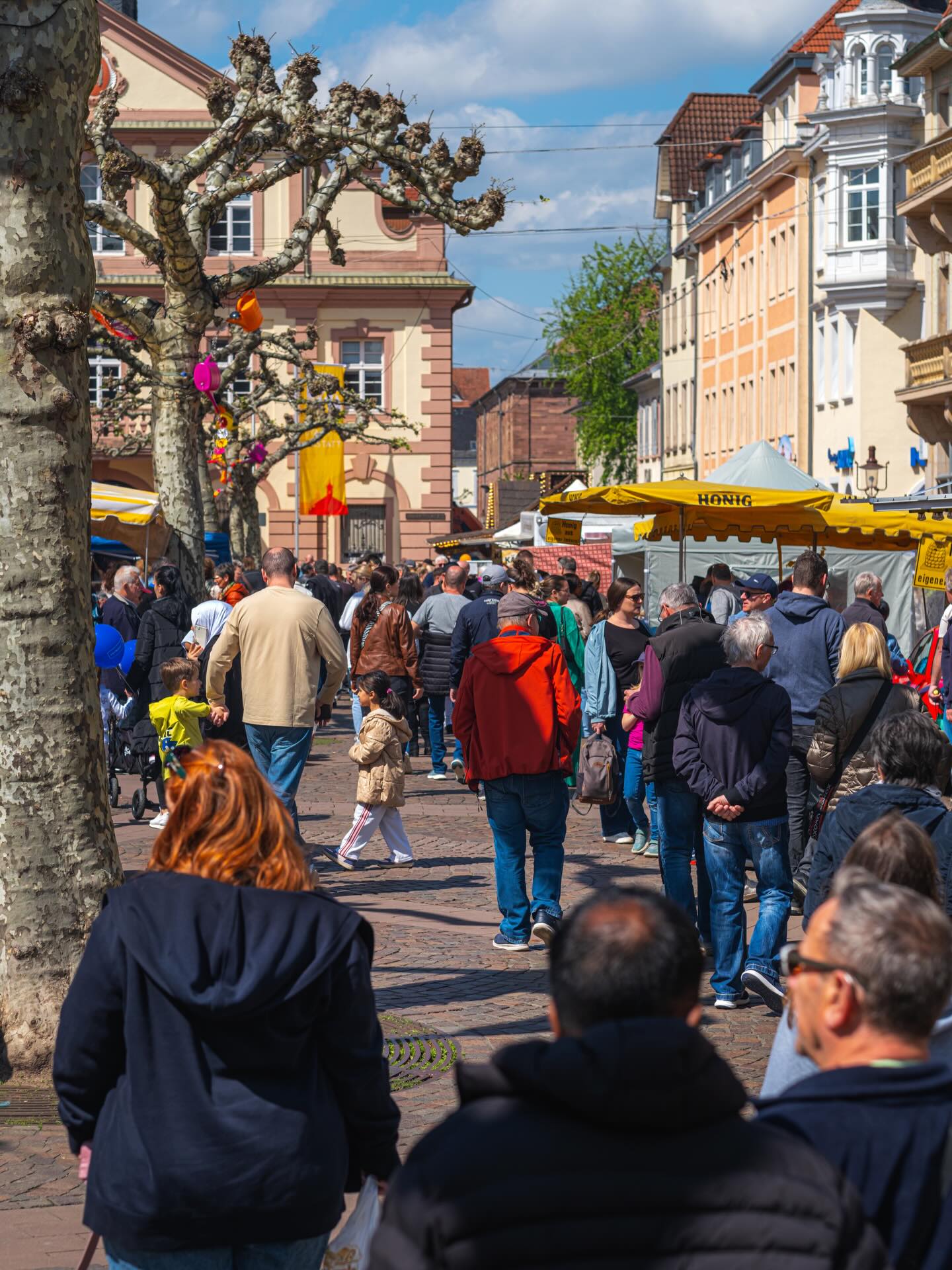Dieser Alternativtext wurde KI-generiert: Eine belebte Straßenszene mit vielen Menschen, die durch einen Marktplatz gehen. Im Hintergrund sind Gebäude mit Architektur zu sehen.