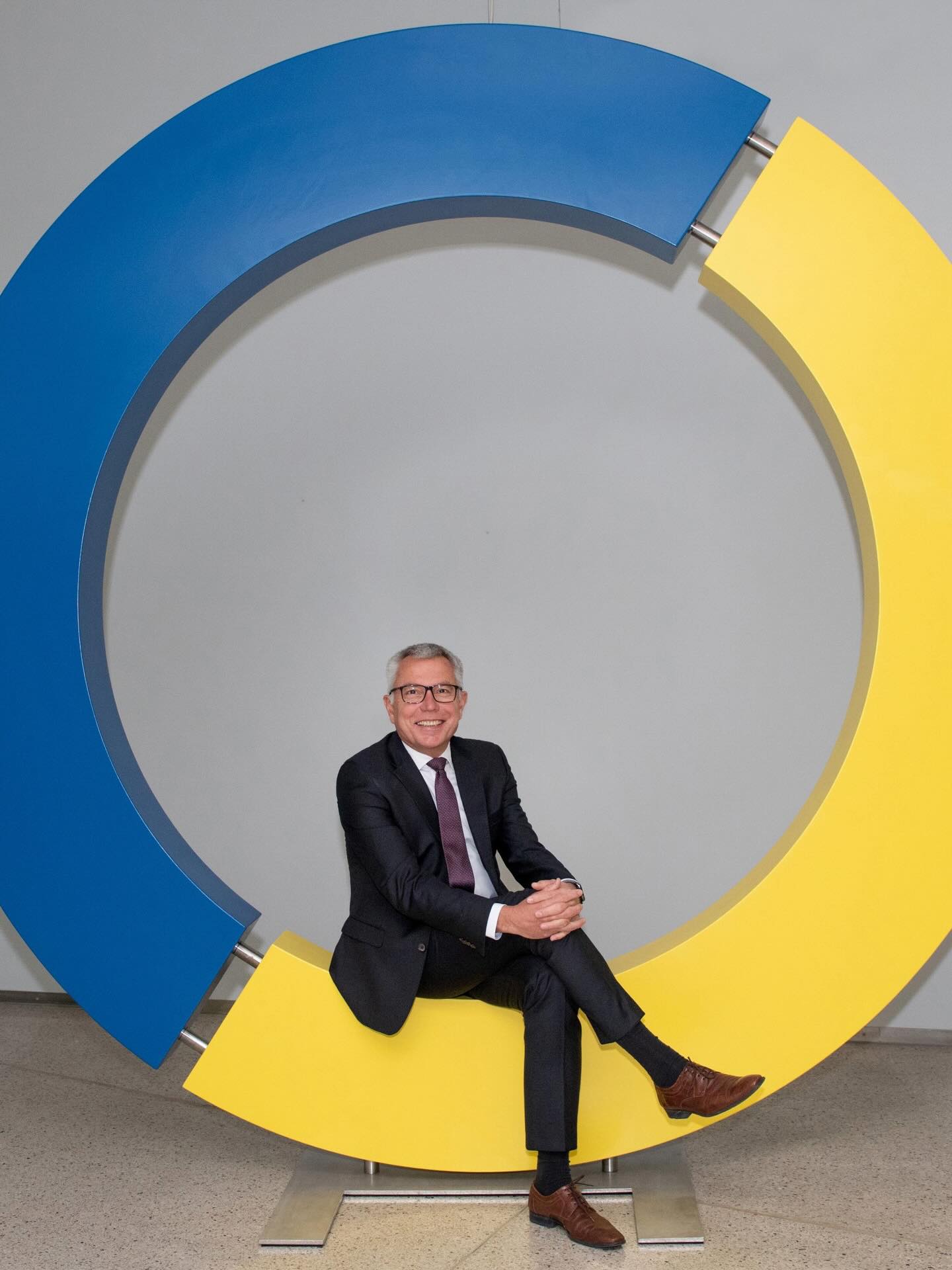 Dieser Alternativtext wurde KI-generiert: Eine Person in einem dunklen Anzug sitzt vor einem großen, farbigen Kreis aus Blau und Gelb. Die Szene ist in einem Innenraum mit hellem Interieur aufgenommen.