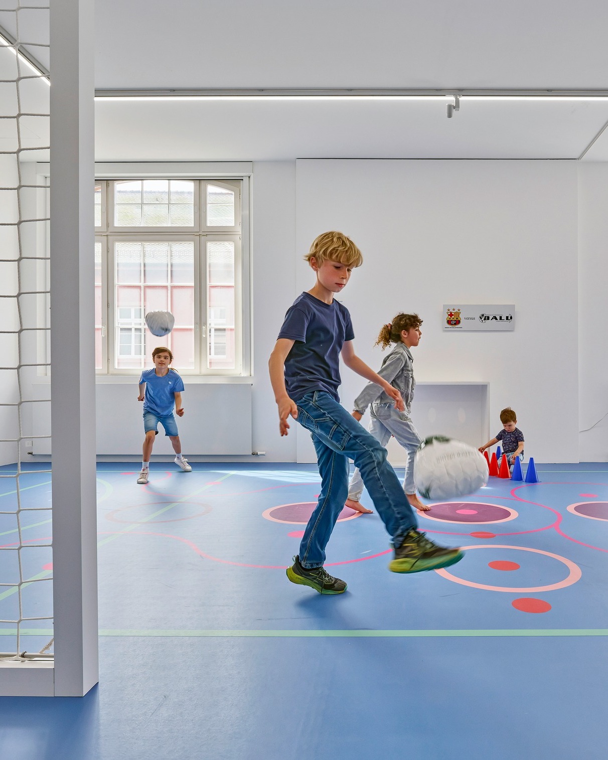 Vier Kinder spielen in einem hellen Raum mit blauem Bodenbelag und bunten Markierungen Fußball. Ein Kind im Vordergrund kickt einen Ball, während ein kleineres Kind im Hintergrund mit roten und blauen Kegeln spielt. An der weißen Wand ist ein Schild mit der Aufschrift „VETUS BALU“ zu sehen. (KI-Alternativtext)