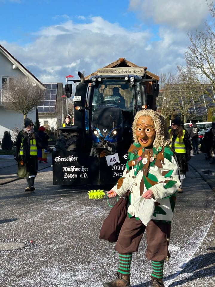 Dieser Alternativtext wurde KI-generiert: Ein farbenfrohes Bild zeigt eine Person in einem aufwändigen, traditionellen Kostüm mit einer Maske vor einem großen Fahrzeug. Im Hintergrund ist ein Gebäude und weitere Personen erkennbar.