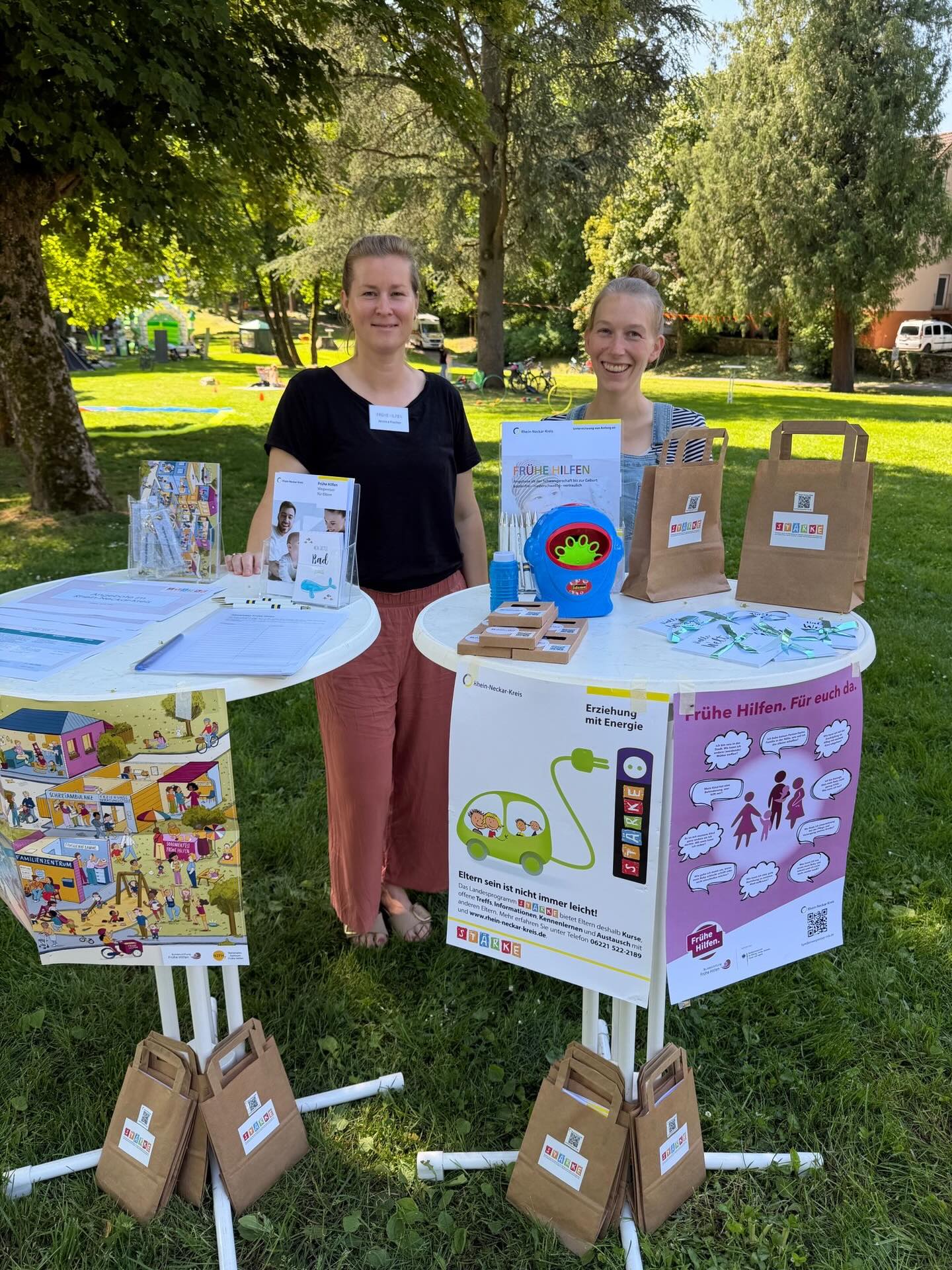 Dieser Alternativtext wurde KI-generiert: Ein Tisch mit Informationsmaterialien und Spielzeug steht im Freien. Mehrere Personen stehen hinter dem Tisch und lächeln.