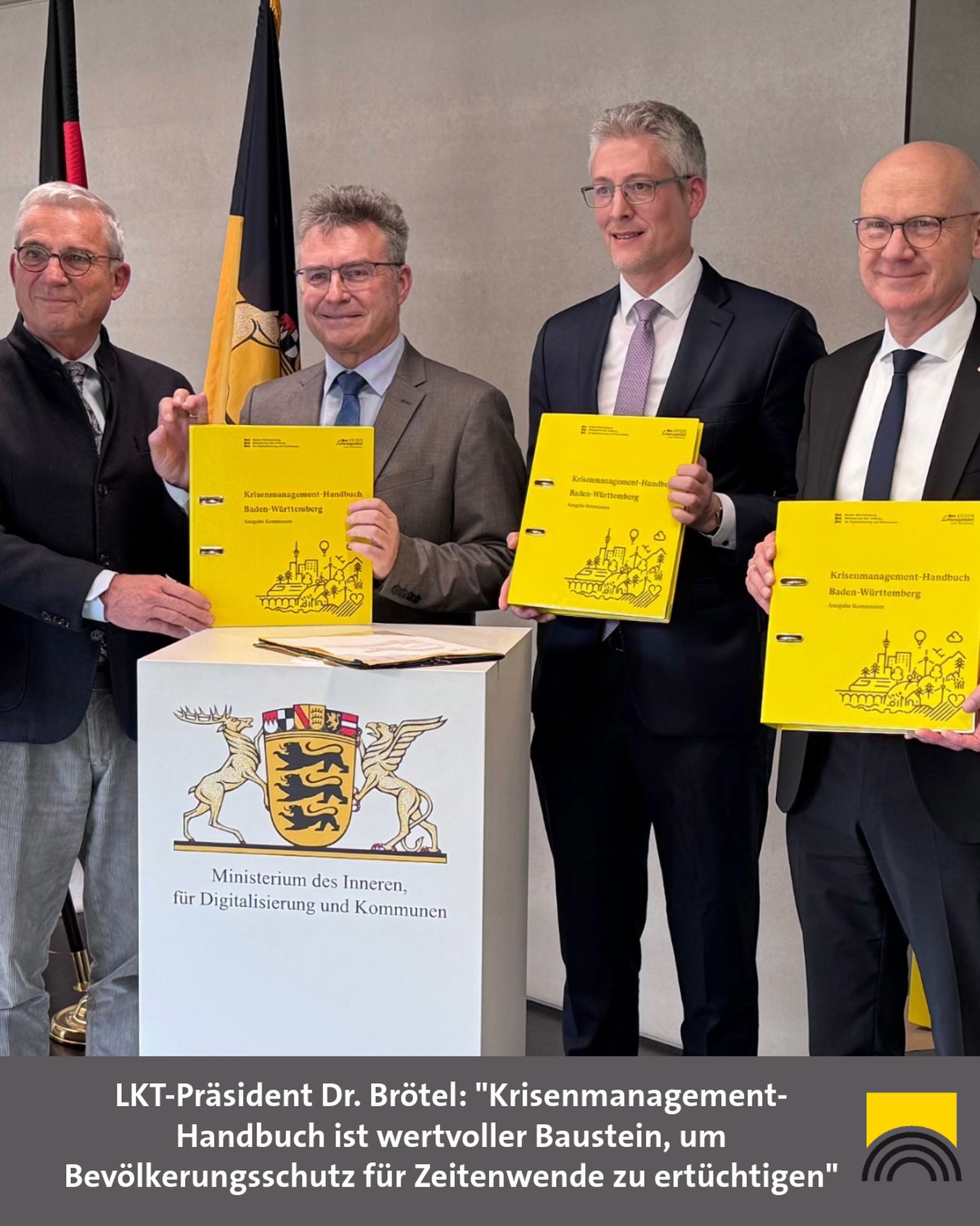 Dieser Alternativtext wurde KI-generiert: Zwei Personen überreichen sich gelbe Mappen mit dem Wappen von Baden-Württemberg. Im Vordergrund liegt ein weiteres Exemplar sowie ein orangefarbener Ordner mit dem Text 'Ministerium des Inneren'.