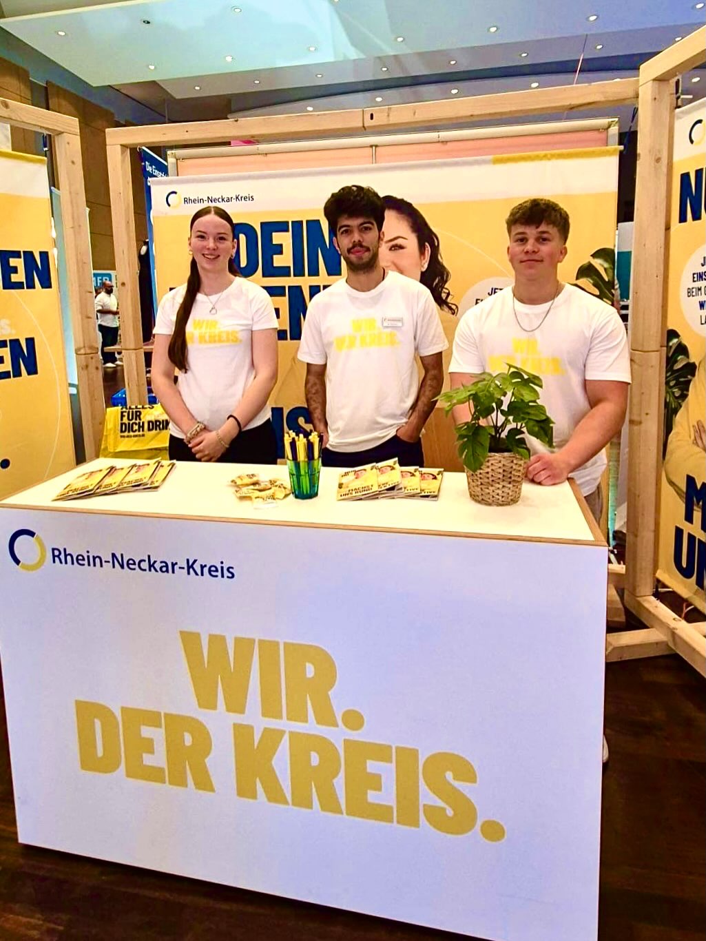 Drei Personen stehen hinter einem weißen Stand mit dem Logo und der Aufschrift „Rhein-Neckar-Kreis“ sowie dem großen Schriftzug „WIR. DER KREIS.“. Alle tragen weiße T-Shirts mit dem gleichen gelben Text. Auf der Theke liegen Broschüren, ein Becher mit Stiften und eine Topfpflanze. (KI-Alternativtext)