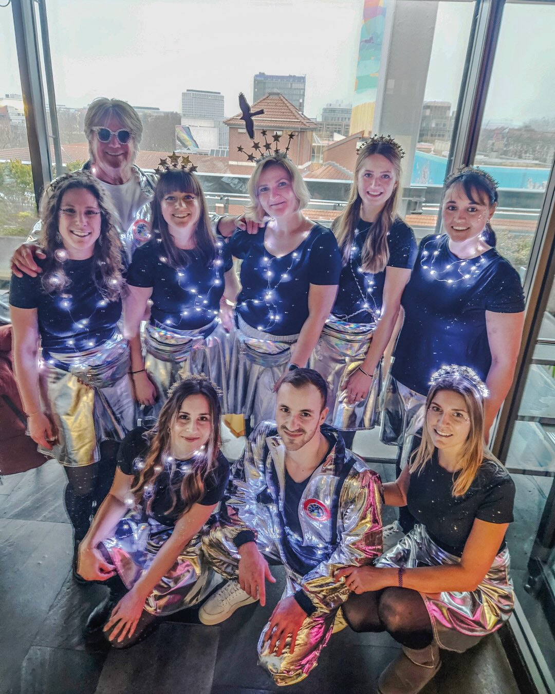 Dieser Alternativtext wurde KI-generiert: Eine Gruppe von Personen in festlicher Kleidung, einige mit Tiara und leuchtenden Accessoires, posiert lächelnd in einem Innenraum mit Blick auf eine Stadtlandschaft. Die Aufnahme ist farbig.