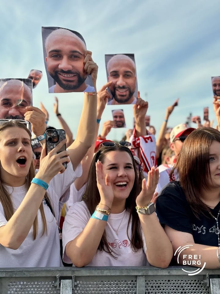 Menschen in einer Menschenmenge halten lächelnde Porträtfotos einer Person in die Höhe, einige rufen oder klatschen. Im Vordergrund sind zwei Personen zu sehen, eine filmt mit dem Smartphone, die andere ruft mit erhobenen Händen. Ein Logo „FREIBURG“ ist unten rechts sichtbar. (KI-Alternativtext)