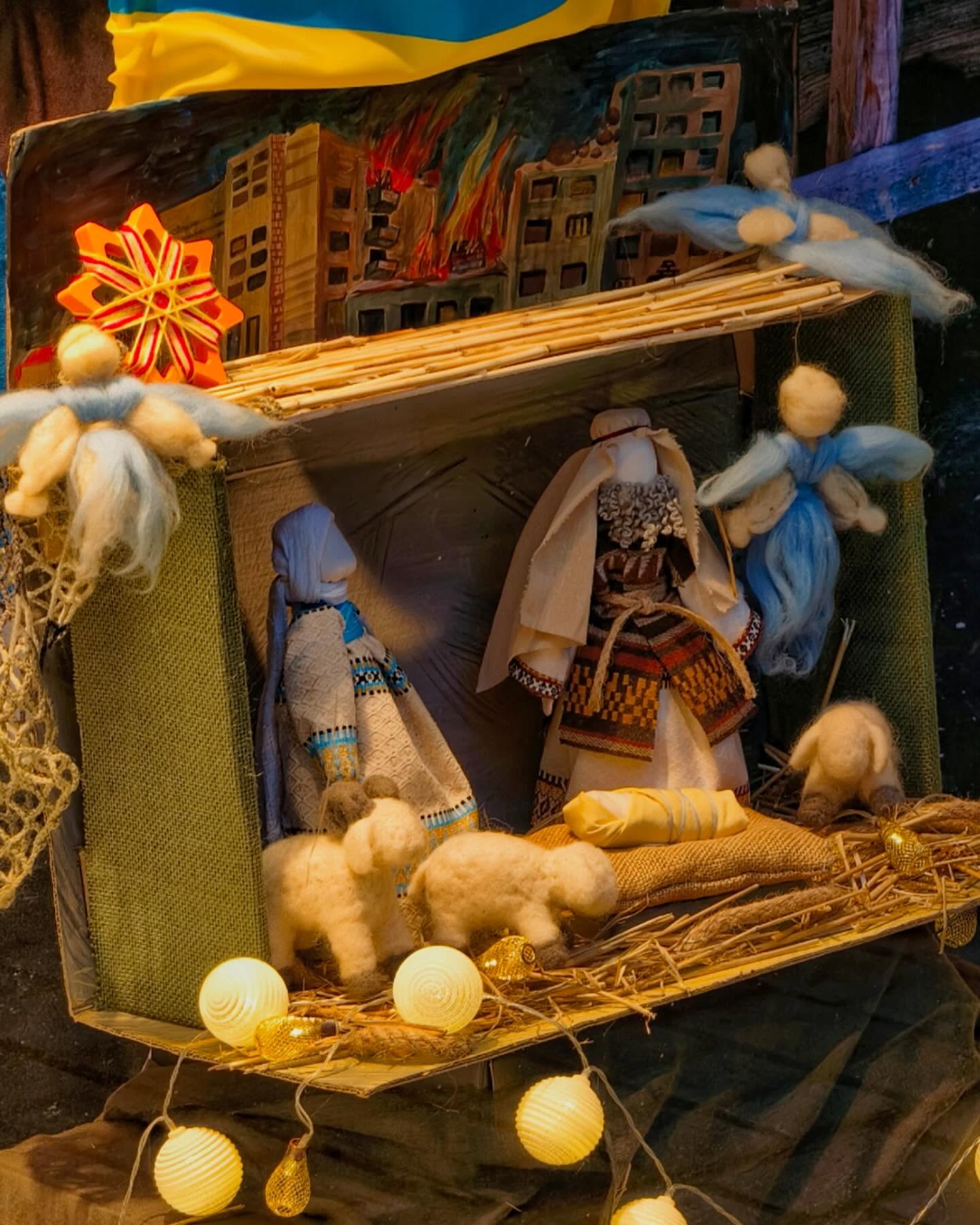 Dieser Alternativtext wurde KI-generiert: Eine Krippendarstellung mit Figuren in einer Stallung. Im Hintergrund sind Gebäude und Lichtquellen zu sehen.