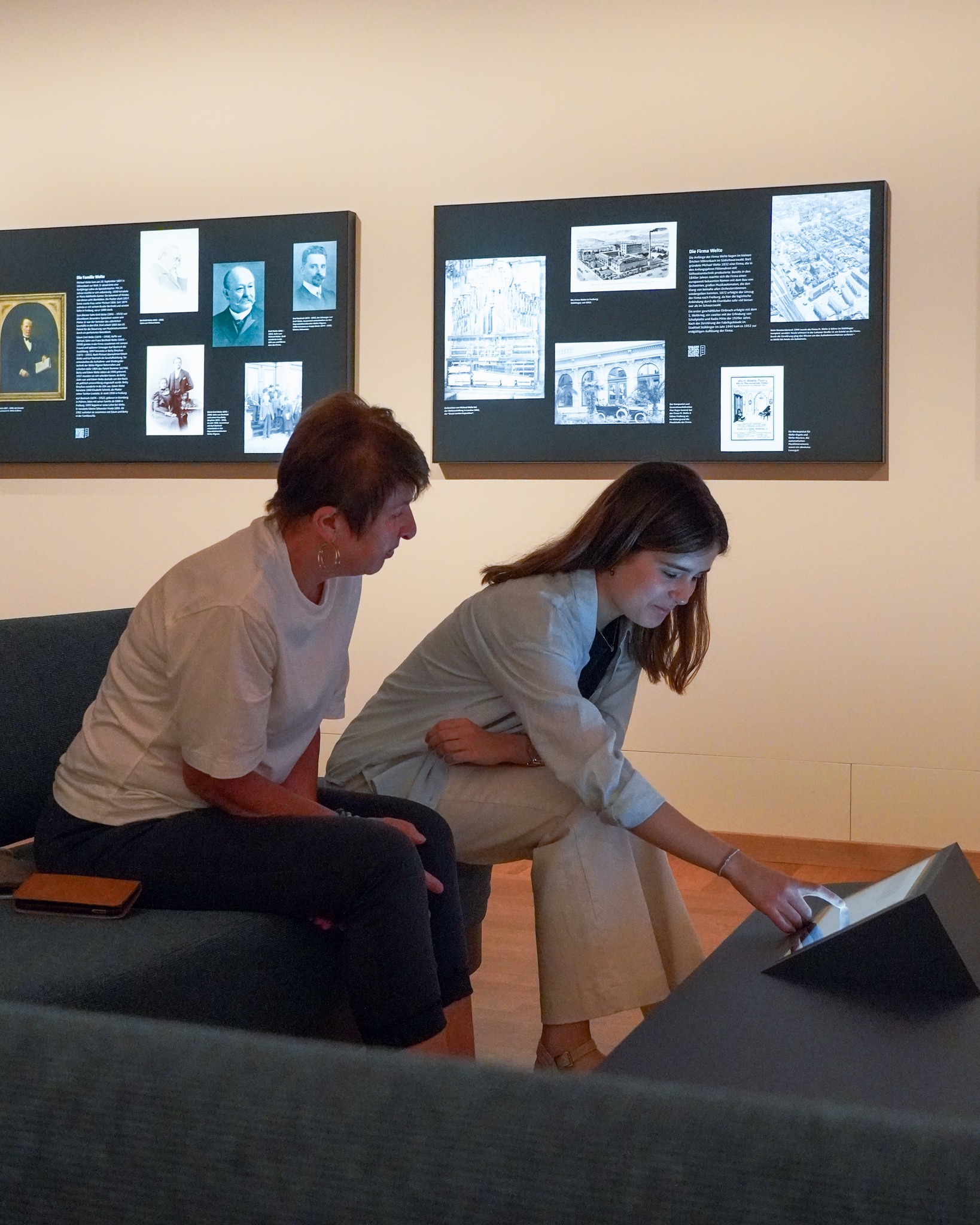 Zwei Personen interagieren in einem Ausstellungsraum mit einem digitalen Angebot. Eine Person sitzt auf einer dunklen Bank, während die andere kniet und ein Display auf einem niedrigen Tisch berührt. Im Hintergrund sind an der Wand zwei große schwarze Infotafeln mit historischen Fotos und Texten zu sehen. (KI-Alternativtext)