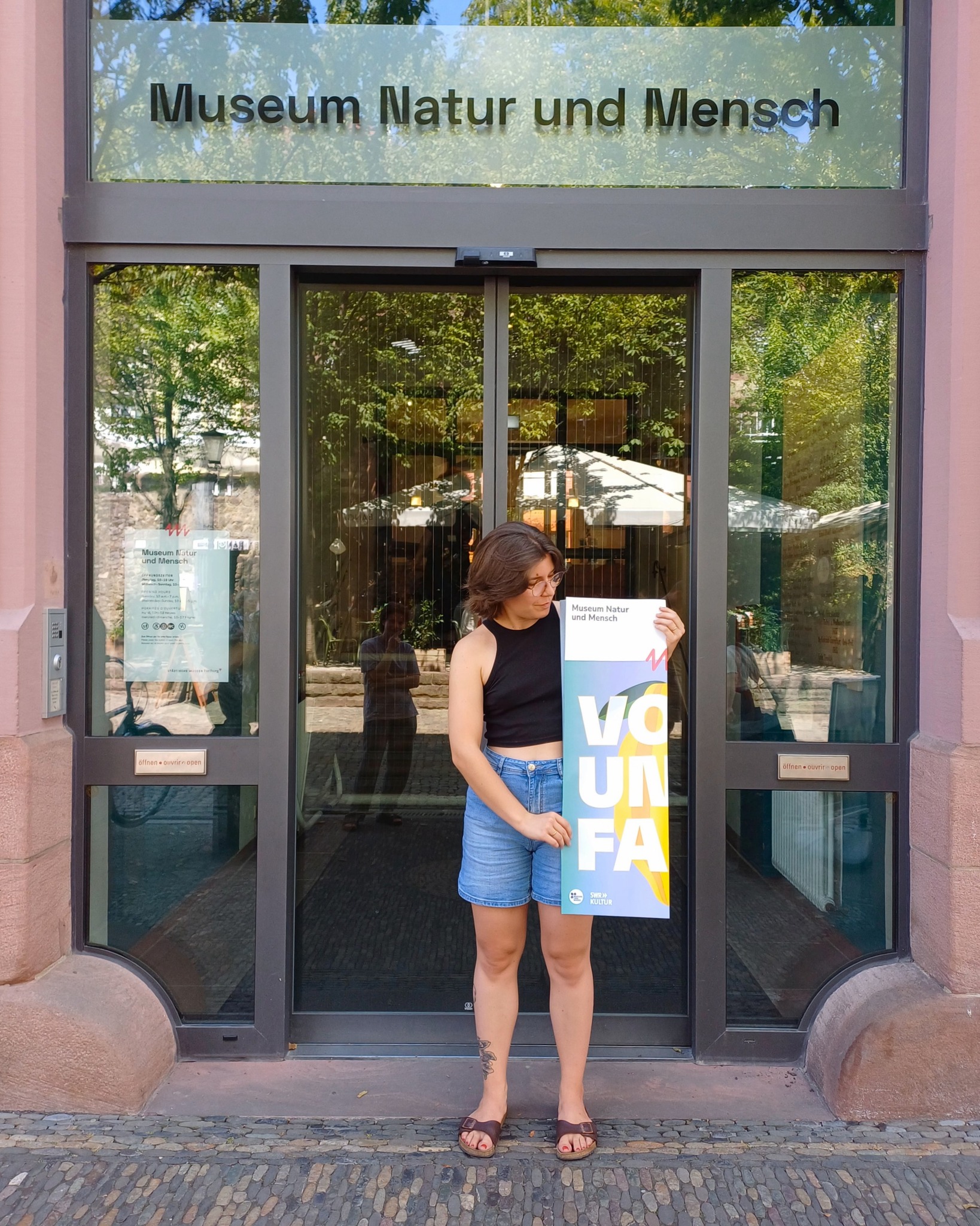 Eine Person steht vor dem Glaseingang des Museum Natur und Mensch. Sie hält ein Plakat, auf dem „Museum Natur und Mensch“, „VOUNFA“ und „SWR KULTUR“ steht. (KI-Alternativtext)