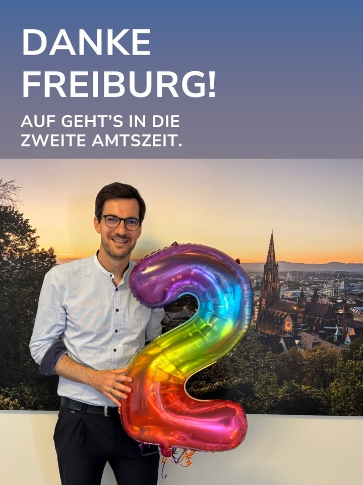 Eine Person mit Brille und hellem Hemd hält einen großen, regenbogenfarbenen Folienballon in Form der Zahl 2 vor einer Stadtansicht mit einem markanten Kirchturm. Oben steht der Text: DANKE FREIBURG! AUF GEHT'S IN DIE ZWEITE AMTSZEIT. (KI-Alternativtext)