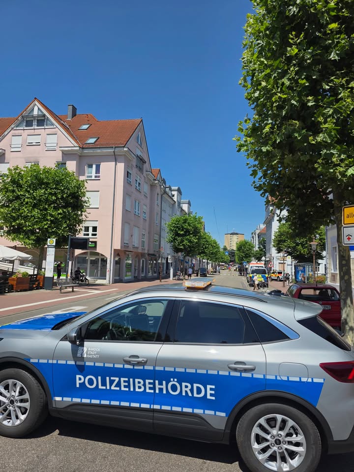 Ein silbernes Polizeifahrzeug mit der Aufschrift „POLIZEIBEHÖRDE“ und „Baden-Württemberg Stuttgart“ steht im Vordergrund auf einer Straße. Im Hintergrund sind weitere Fahrzeuge, darunter ein Einsatzwagen, sowie Personen auf dem Gehweg vor mehrstöckigen Gebäuden und Bäumen zu sehen. (KI-Alternativtext)