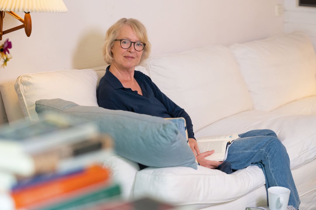 Eine Person mit kurzen blonden Haaren und Brille sitzt auf einem weißen Sofa und hält ein offenes Buch auf dem Schoß. Sie trägt ein dunkelblaues Hemd und Jeans. Im unscharfen Vordergrund liegt ein Stapel Bücher. (KI-Alternativtext)
