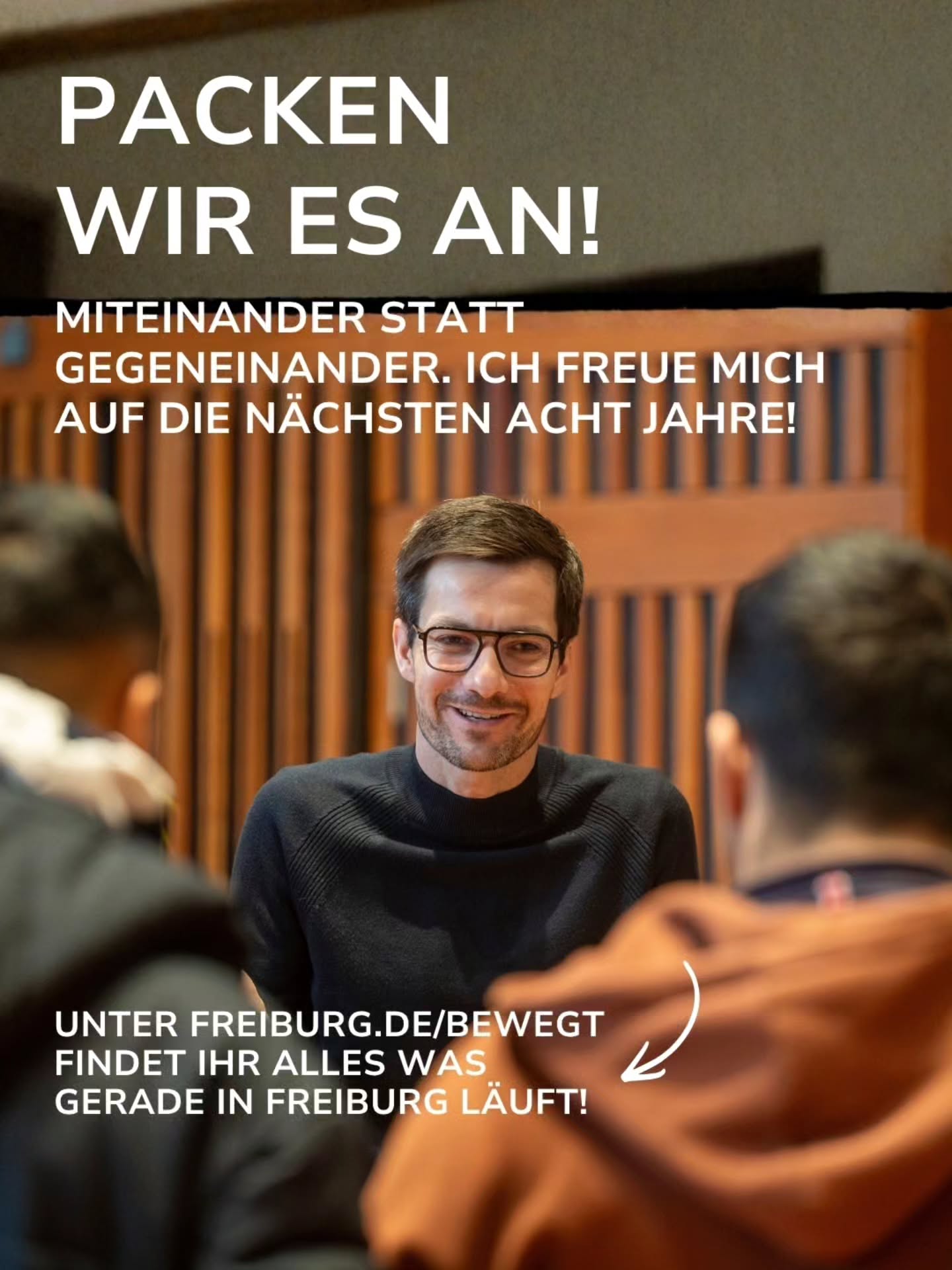 Eine Person mit Brille und dunklem Pullover sitzt lächelnd an einem Tisch und blickt in die Kamera, während im Vordergrund zwei weitere Personen von hinten zu sehen sind. Der Text im Bild lautet: „PACKEN WIR ES AN! MITEINANDER STATT GEGENEINANDER. ICH FREUE MICH AUF DIE NÄCHSTEN ACHT JAHRE!“ Unten ergänzt ein Text: „UNTER FREIBURG.DE/BEWEGT FINDET IHR ALLES WAS GERADE IN FREIBURG LÄUFT!“ (KI-Alternativtext)
