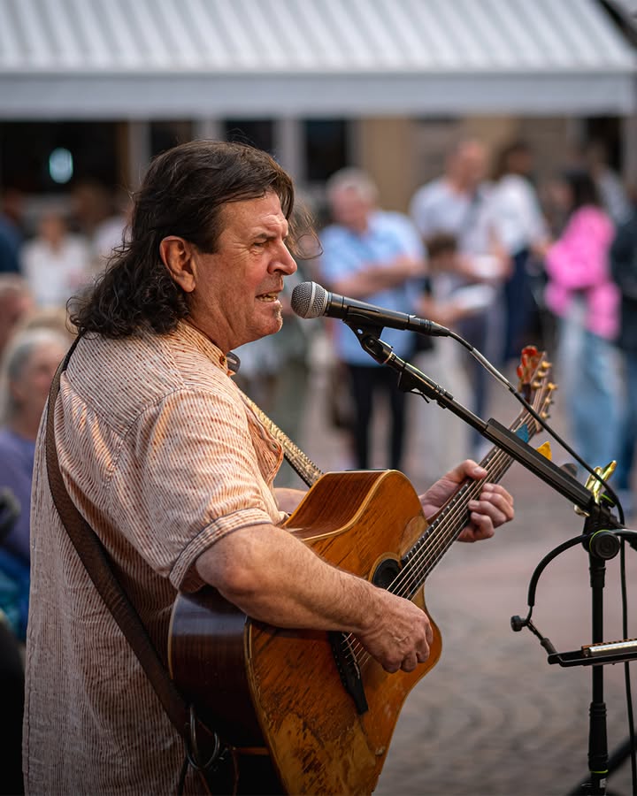Eine Person mit langem dunklem Haar und gestreiftem Hemd spielt eine akustische Gitarre und singt in ein Mikrofon. Im Hintergrund ist eine unscharfe Menschenmenge vor einem weißen Zelt zu sehen. (KI-Alternativtext)
