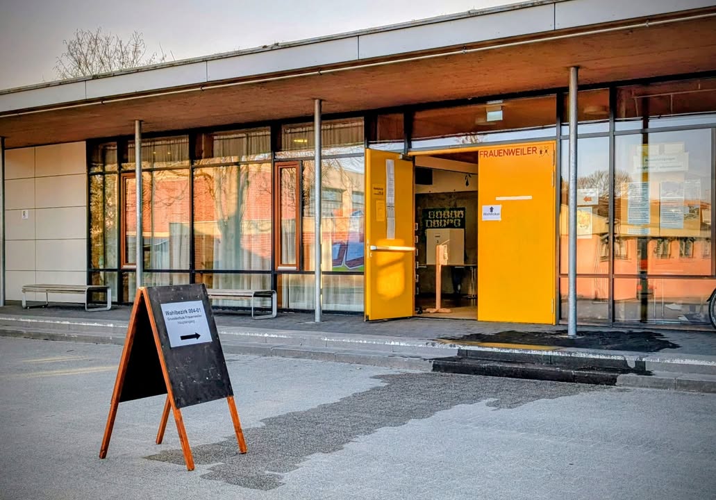 Dieser Alternativtext wurde KI-generiert: Ein Gebäude mit einer großen Glasfassade und mehreren Türen. Vor dem Eingang steht ein kleines Schild auf einem Gestell.