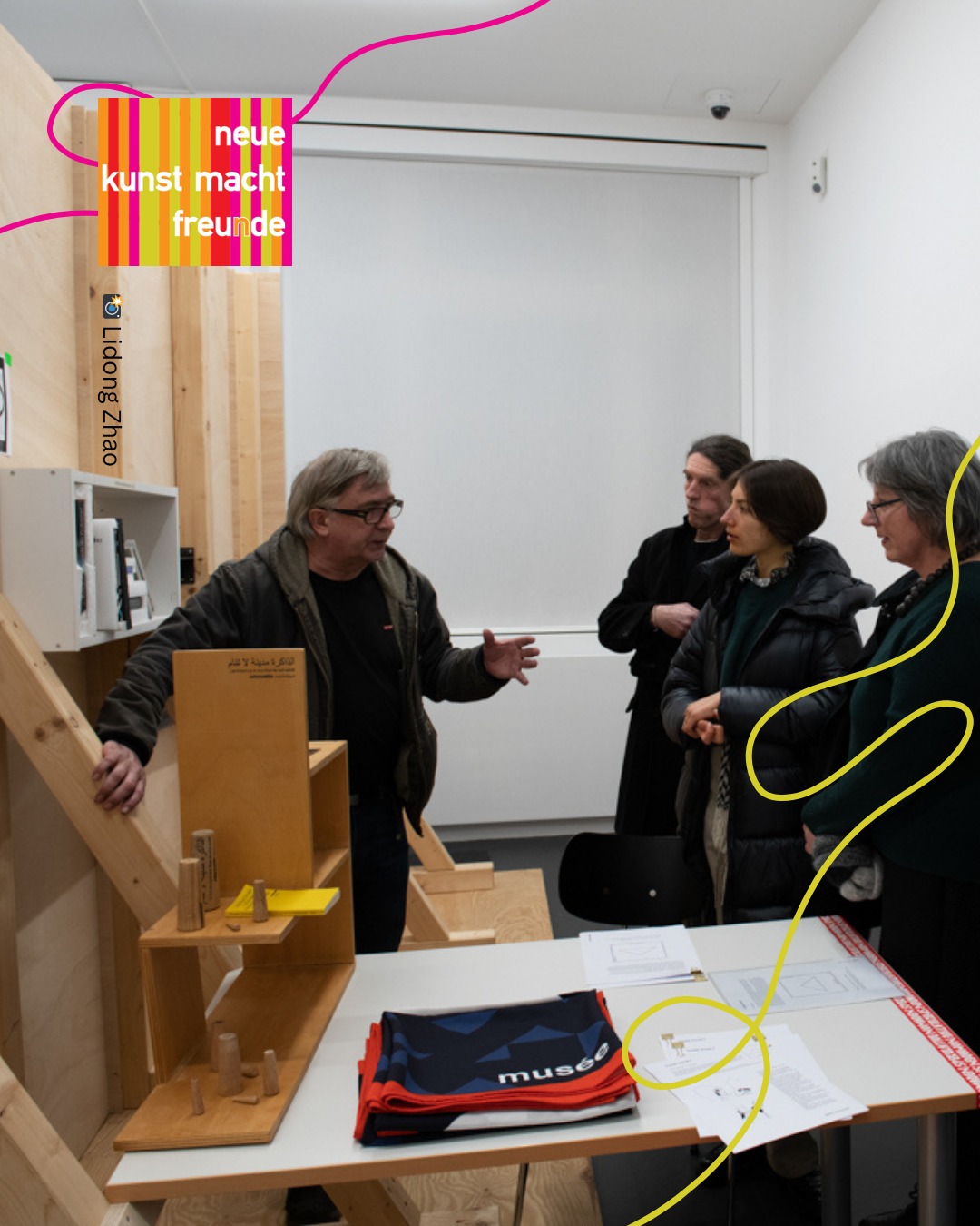 Eine Person mit Brille erklärt vor einer Gruppe von drei Zuhörern ein Holzmodell auf einem Tisch; im Hintergrund ein farbiges Schild mit dem Text „neue kunst macht freunde“. Auf dem Tisch liegen auch gefaltete Taschen mit dem Aufdruck „musée“. (KI-Alternativtext)