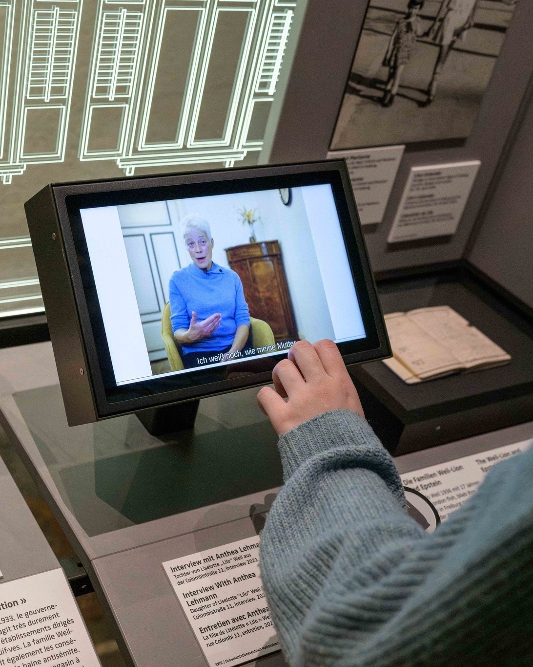 Eine Hand bedient einen Touchscreen-Monitor in einer Ausstellung. Auf dem Display ist ein Video-Interview mit einer Person mit kurzen weißen Haaren und blauem Pullover zu sehen, dazu die Untertitel "Ich weiß noch, wie meine Mutter". Der Sockel trägt die Beschriftung "Interview mit Anthea Lehmann" in drei Sprachen. (KI-Alternativtext)
