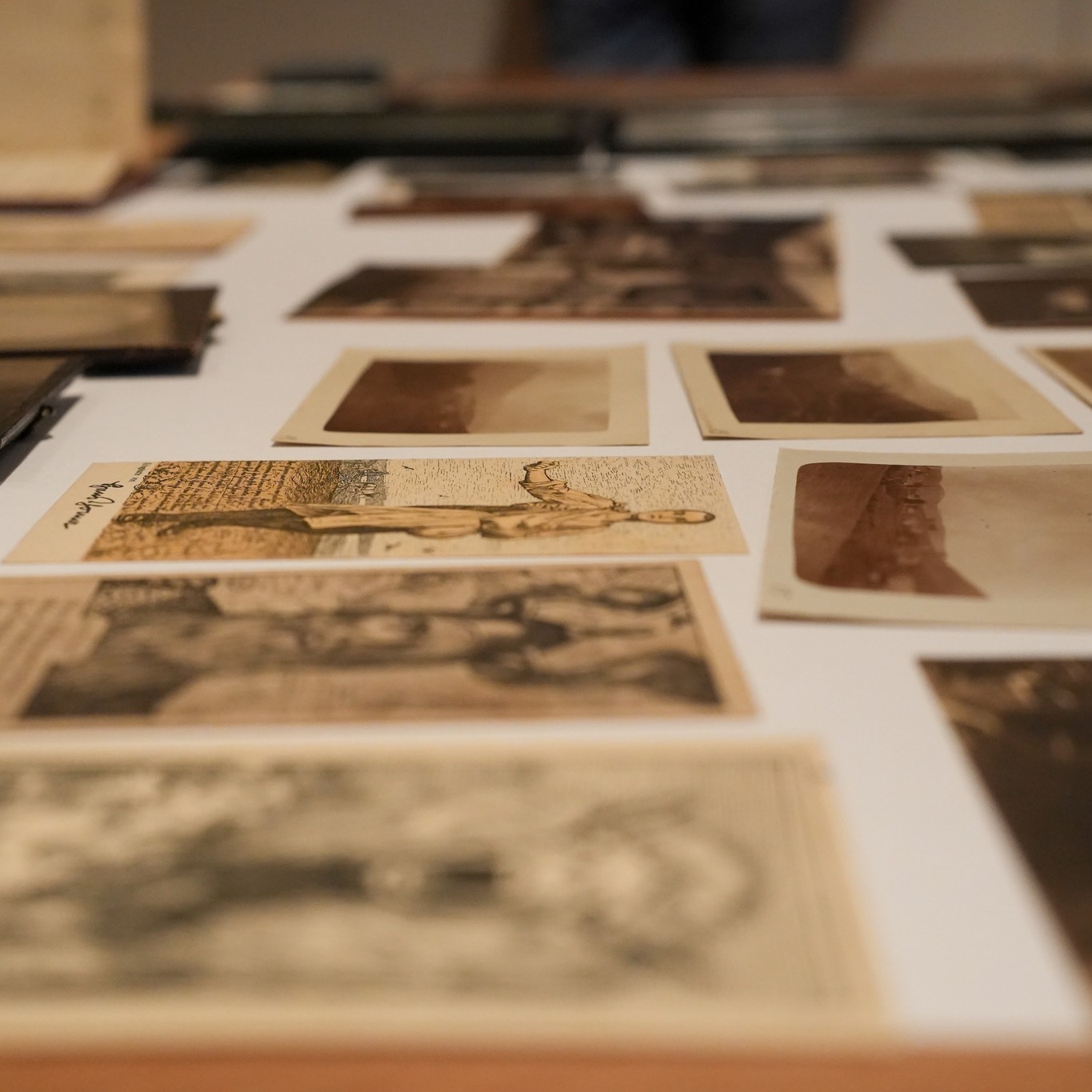 Auf einer weißen Unterlage sind zahlreiche alte, sepiafarbene Fotografien und Postkarten ausgebreitet. Eine Postkarte im mittleren Bereich ist scharf abgebildet und zeigt eine stehende Person in einer Landschaft mit Text links daneben. Die übrigen Bilder im Vordergrund und Hintergrund sind unscharf. (KI-Alternativtext)