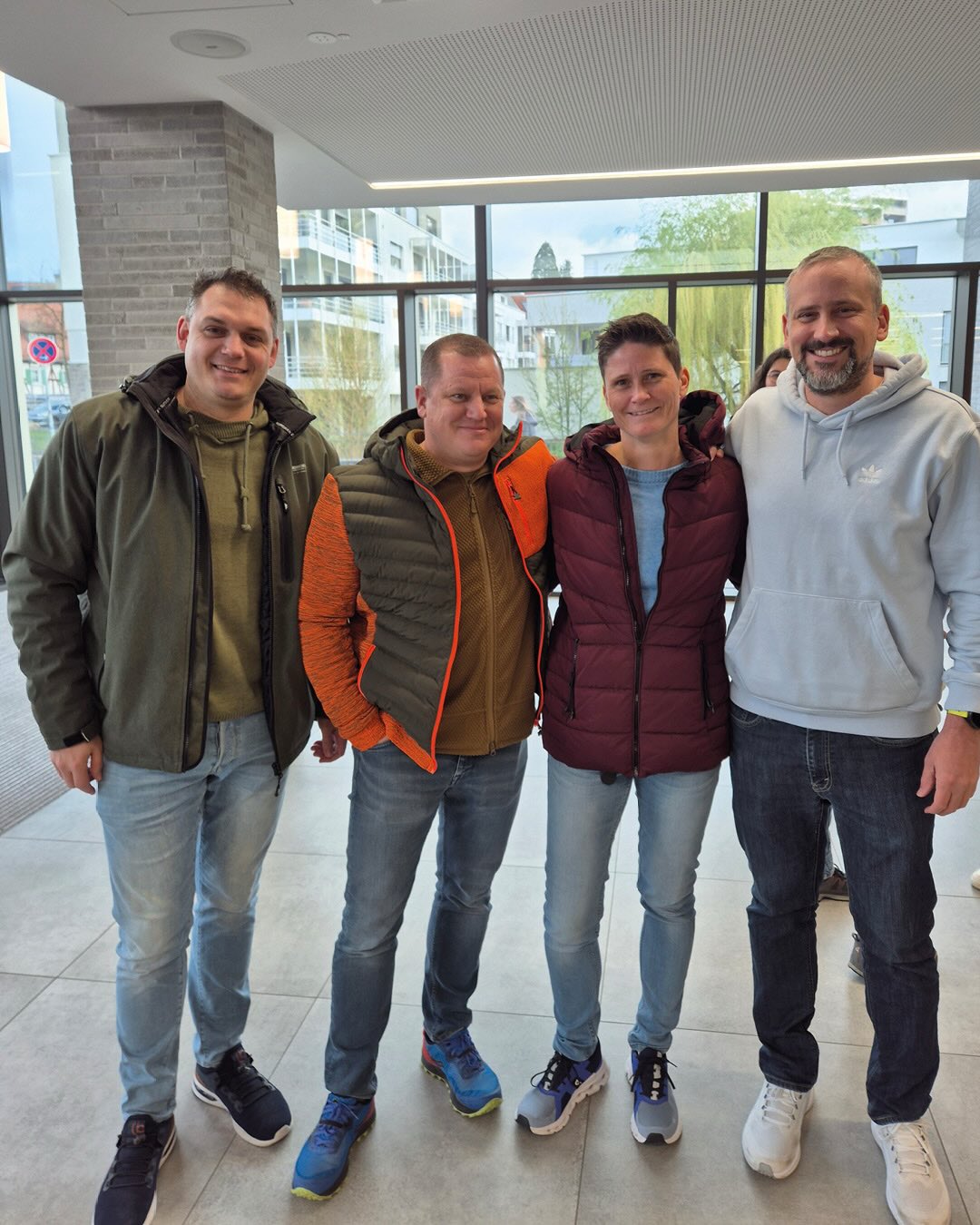Dieser Alternativtext wurde KI-generiert: Eine Aufnahme von vier Personen, die nebeneinander stehen und lächeln. Sie befinden sich in einem Innenraum mit Fensterfront, durch die Gebäude im Hintergrund zu sehen sind.