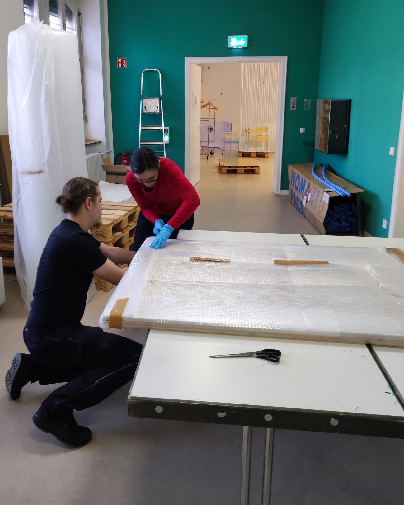 Dieser Alternativtext wurde KI-generiert: Zwei Personen arbeiten in einem Raum mit einer hellen, türkisfarbenen Wand. Sie scheinen ein großes, in Luftpolsterfolie verpacktes Objekt zu handhaben.