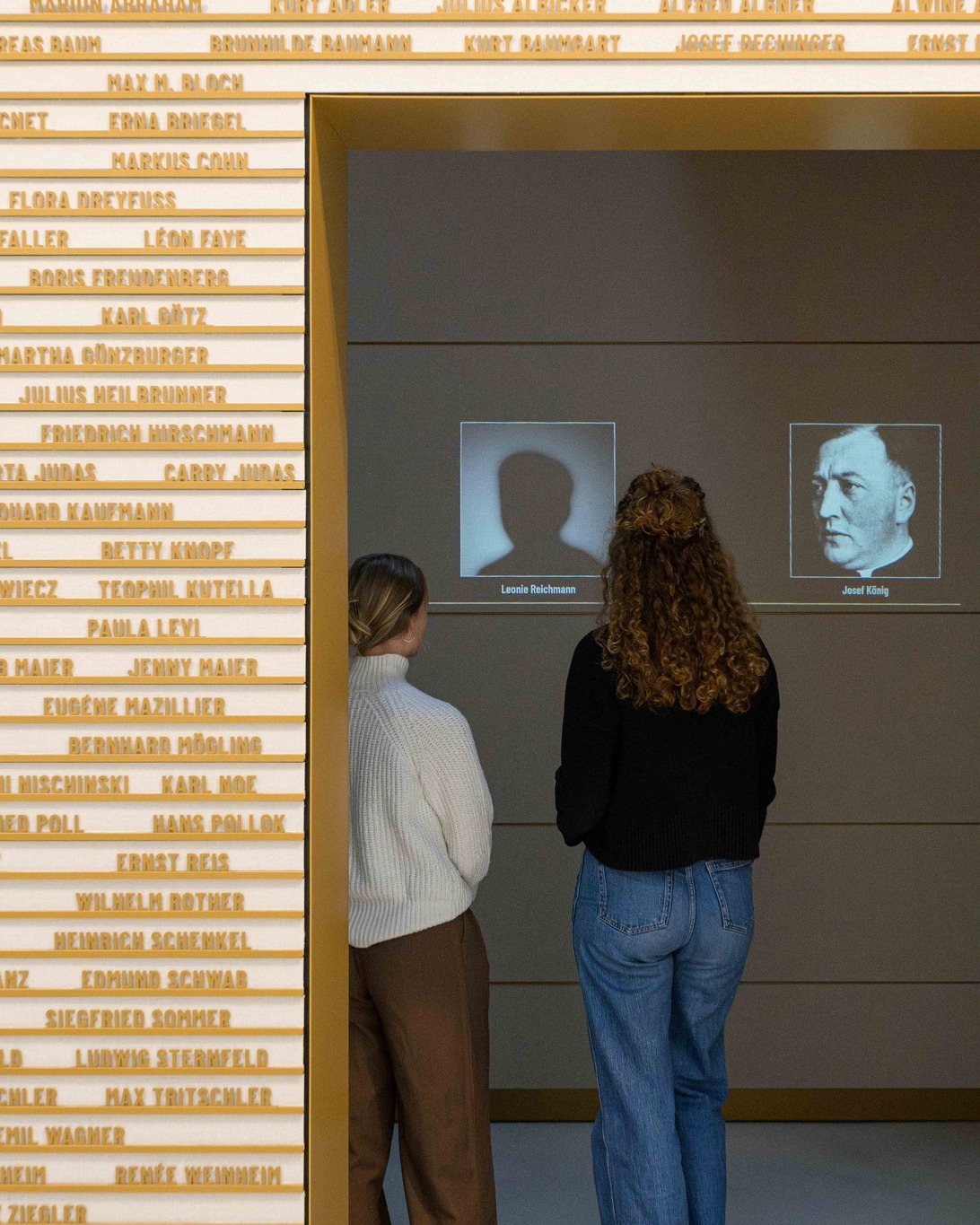 Zwei Personen stehen mit dem Rücken zur Kamera und betrachten eine Ausstellung. Links ist eine Wand mit zahlreichen Namen in Goldbuchstaben zu sehen. Rechts zeigt ein Bildschirm eine Silhouette mit der Beschriftung „Leonie Reichmann“ neben einem Porträtfoto eines Mannes mit der Beschriftung „Josef König“. (KI-Alternativtext)