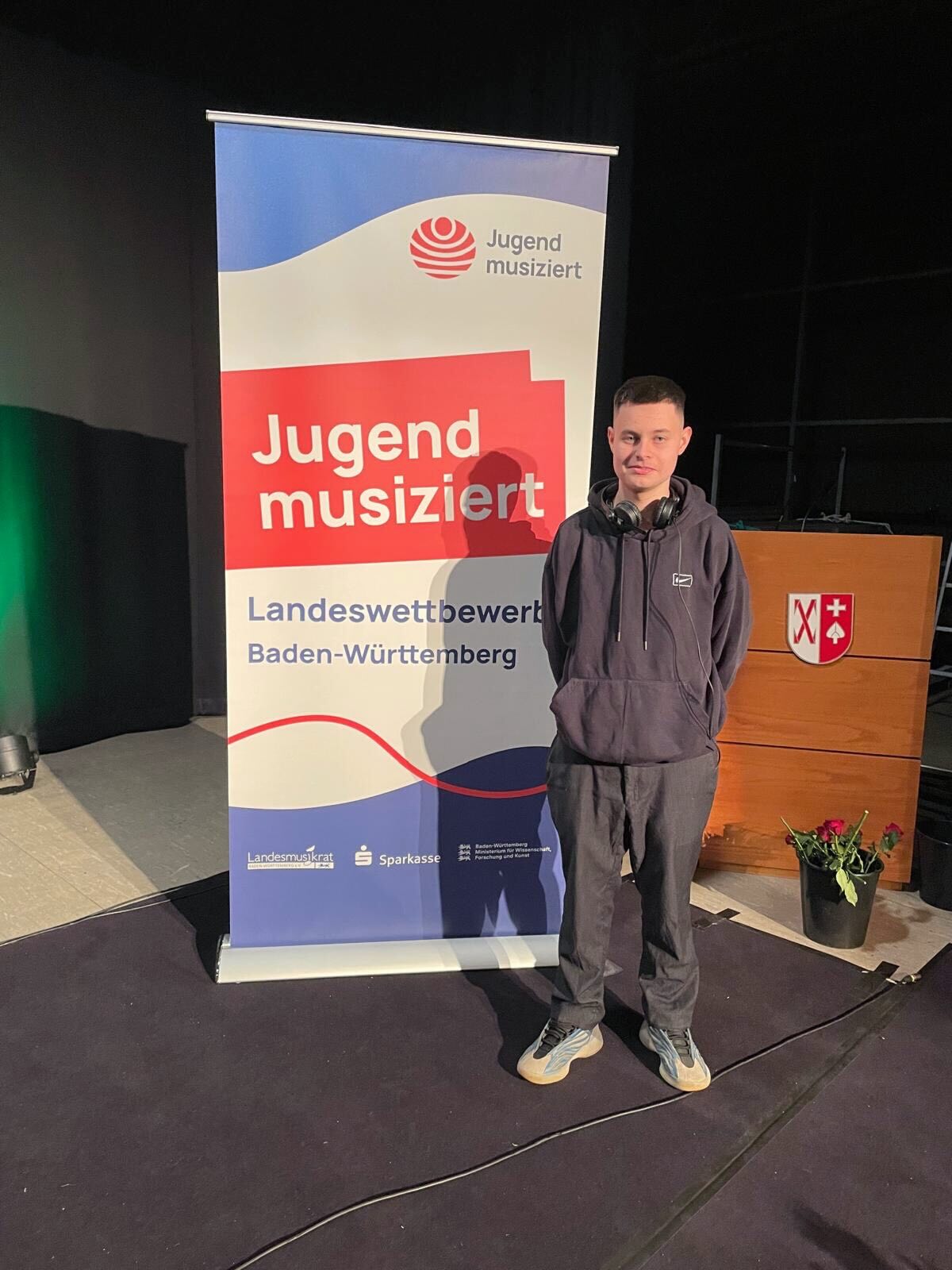 Dieser Alternativtext wurde KI-generiert: Eine Person steht vor einem Banner mit Text und Logos. Das Bild ist farbig und zeigt eine Indoor-Szene mit Bühnenbeleuchtung.