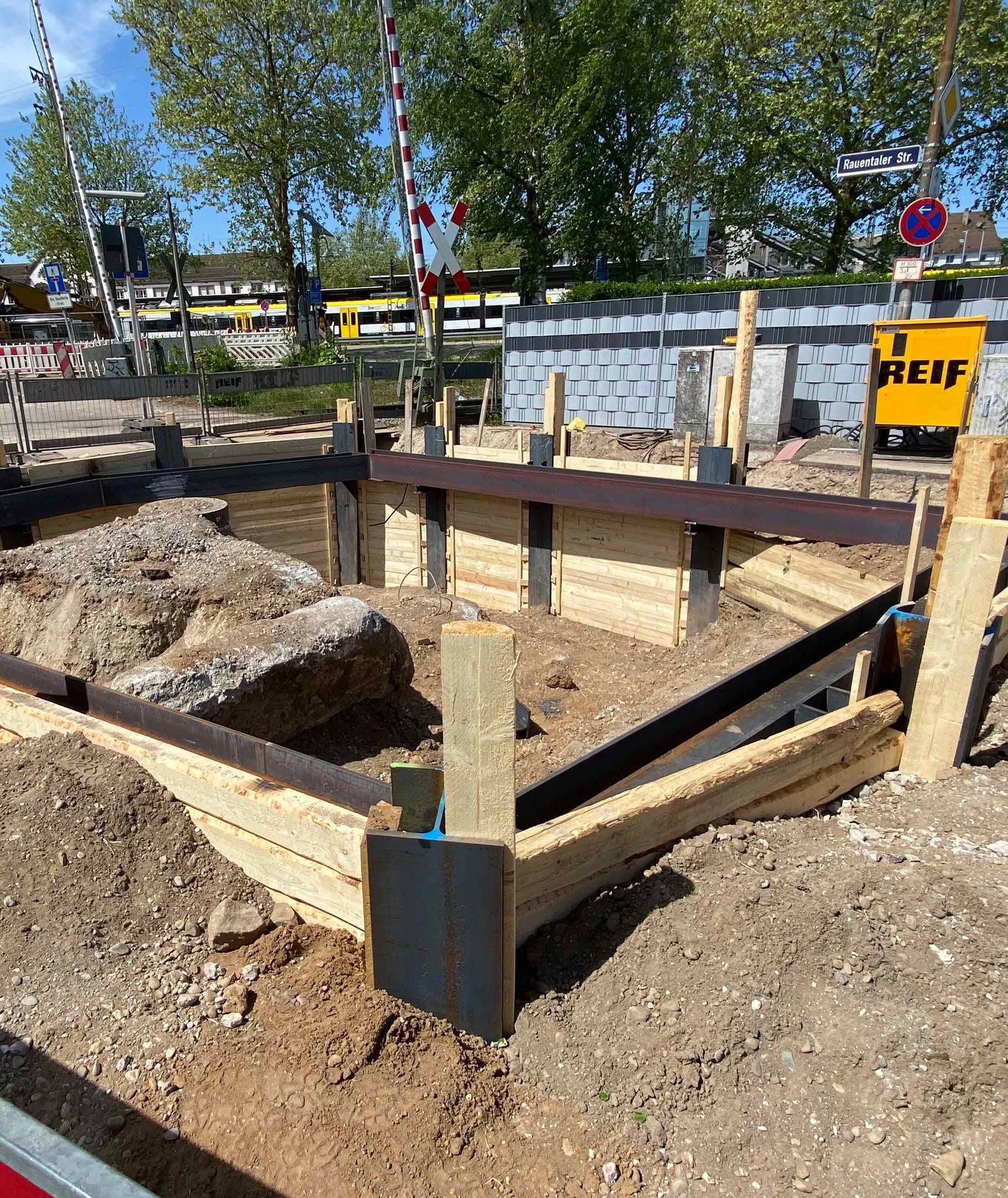 Dieser Alternativtext wurde KI-generiert: Eine Baustelle mit einem Fundament in Bau. Im Hintergrund sind eine Straße, ein Schranke und Gebäude zu sehen.