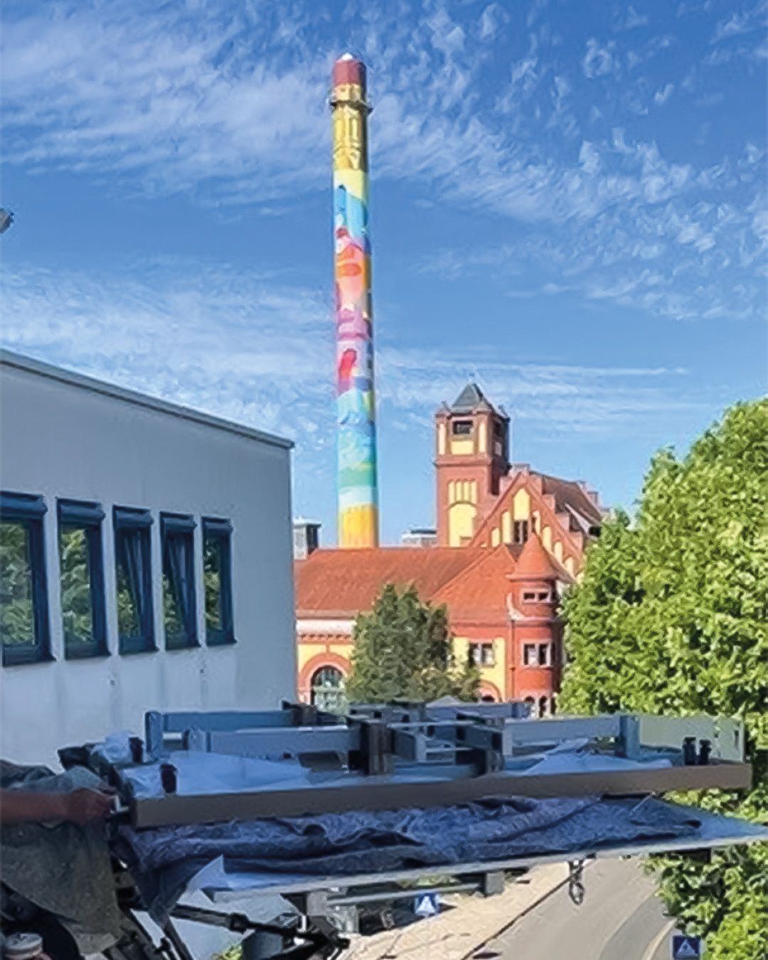 Dieser Alternativtext wurde KI-generiert: Ein hoher Schornstein mit einem farbenfrohen Kunstwerk an der Seite erstreckt sich in den blauen Himmel. Im Hintergrund sind Gebäude und Bäume zu erkennen.