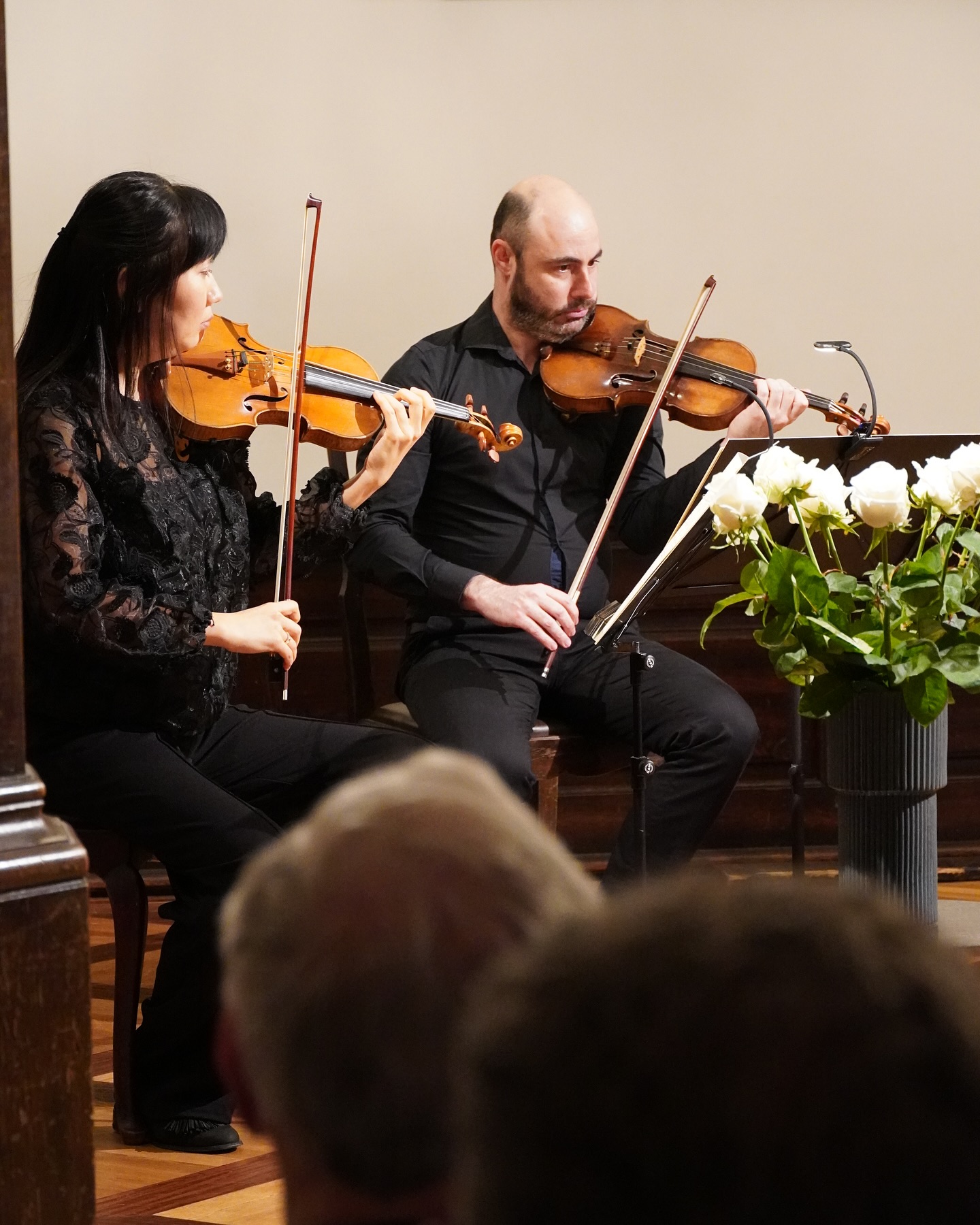 Zwei Musiker spielen Violine. Sie sitzen auf Stühlen vor einem Notenständer mit weißen Rosen.