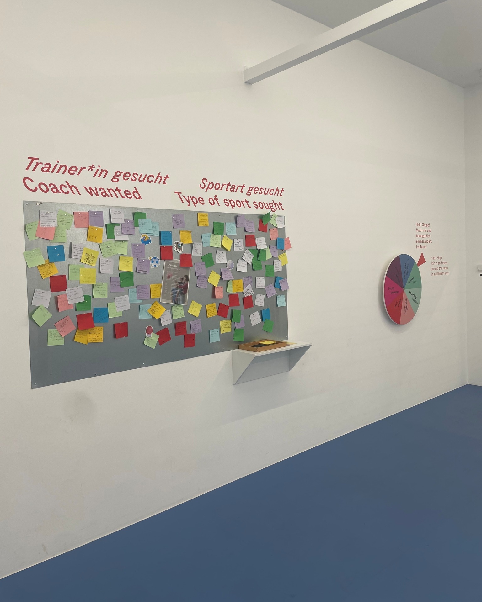 Weiße Wand mit grauer Pinnwand voller bunter Haftnotizen und einem kleinen Regal darunter. Oben steht in Rot „Trainer*in gesucht Coach wanted“ und „Sportart gesucht Type of sport sought“. Rechts hängt ein buntes Rad mit dem Text „Halt! Stopp! Mach mit und bewege dich einmal anders im Raum!“. (KI-Alternativtext)