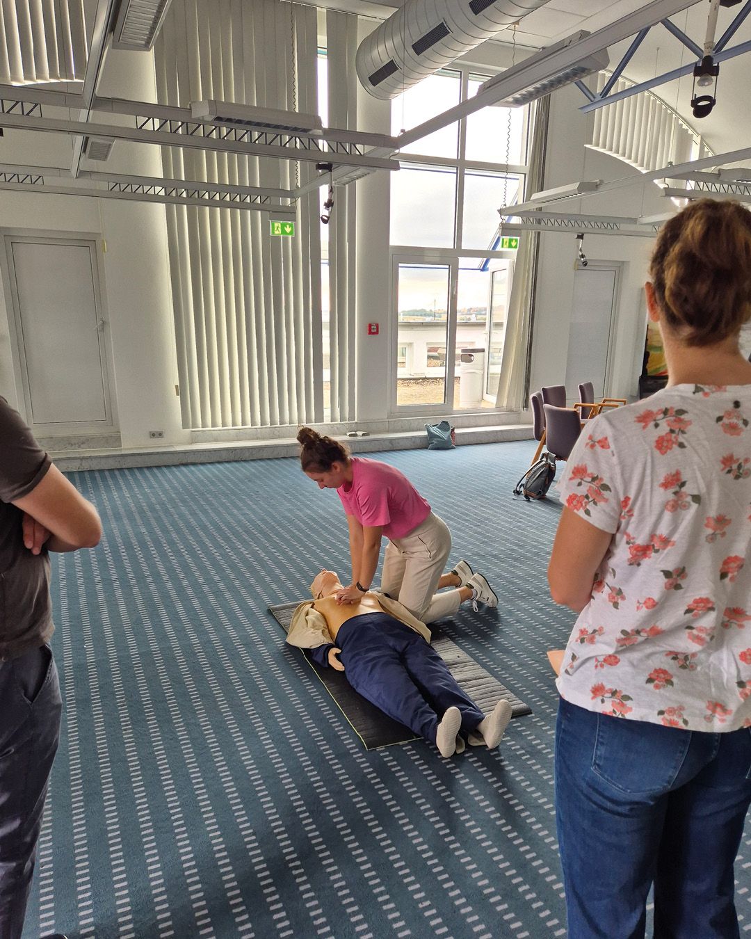 Eine Person kniet auf einer Matte und übt Herzdruckmassage an einer Reanimationspuppe. Zwei weitere Personen stehen im Raum und beobachten die Übung. (KI-Alternativtext)