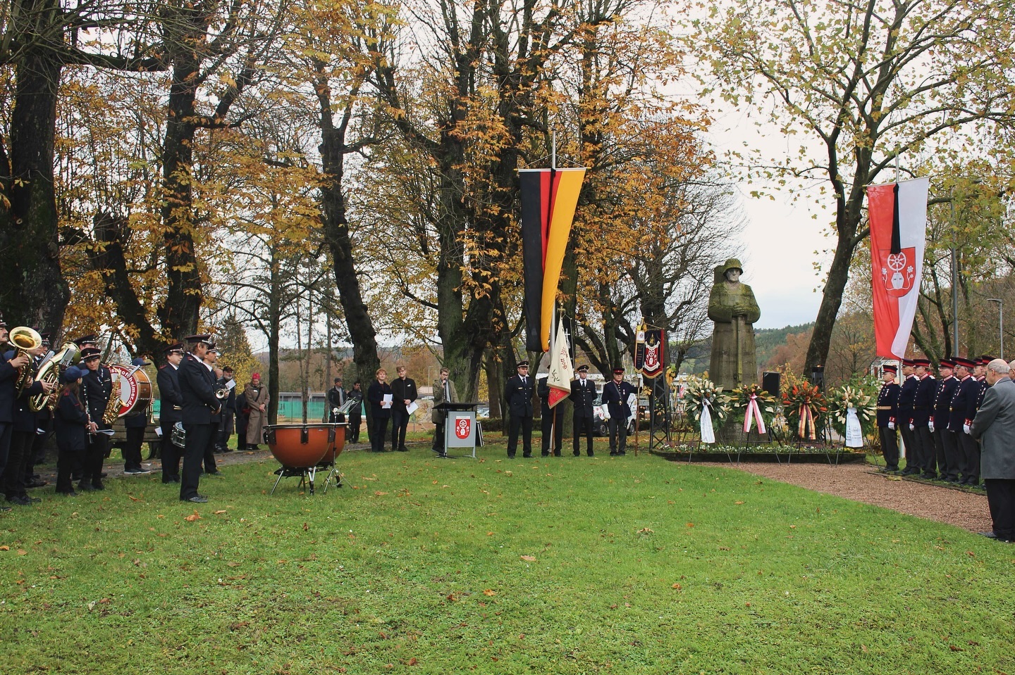Eine Zeremonie findet im Freien unter Bäumen statt. Musiker*innen spielen, Menschen stehen in Formationen und Kränze liegen vor einer Statue.
