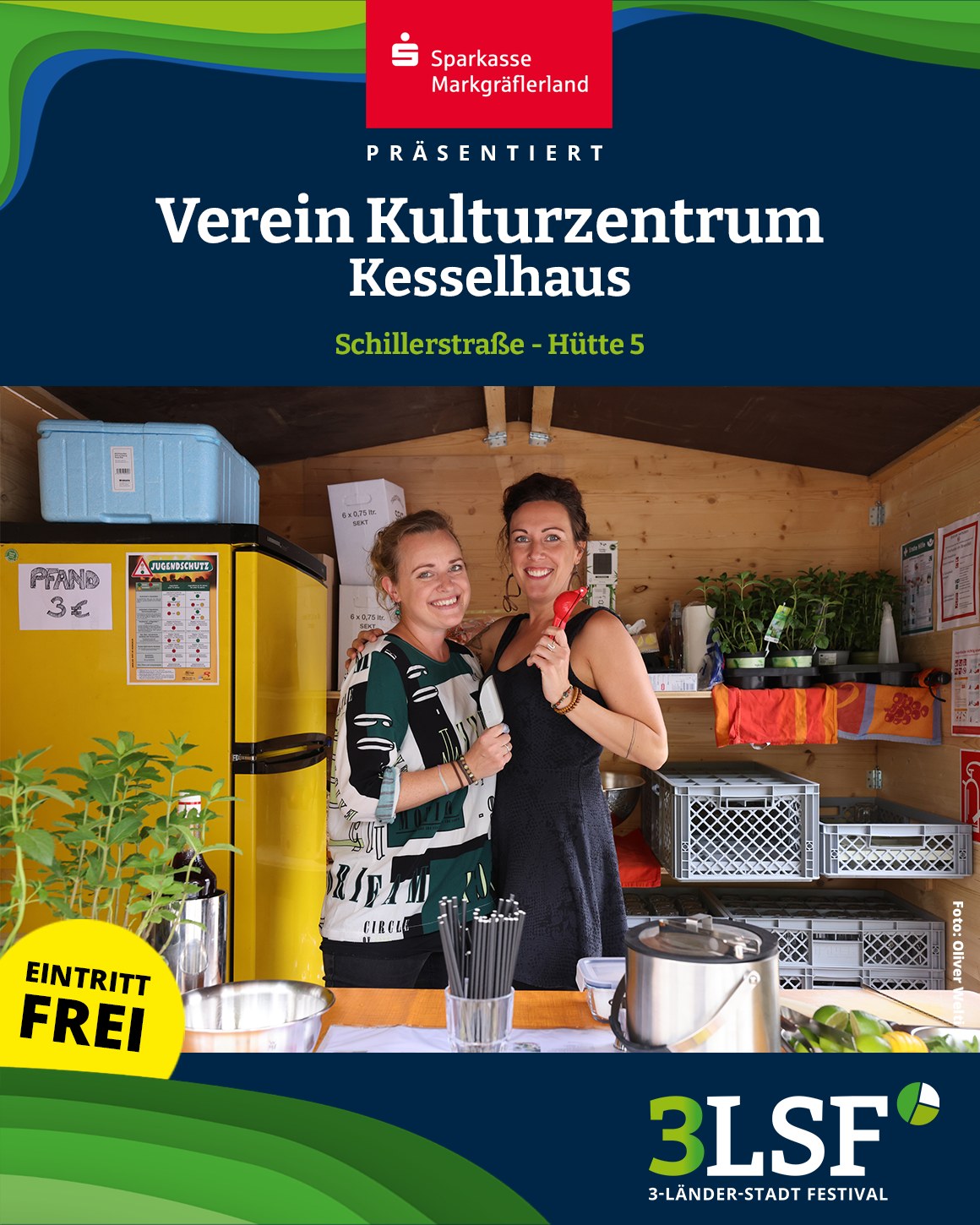 Dieser Alternativtext wurde KI-generiert: Ein farbiges Bild zeigt zwei lächelnde Personen vor einem Stand mit Pflanzen und Getränken. Im Hintergrund ist ein Gebäude mit einem Dach zu sehen.