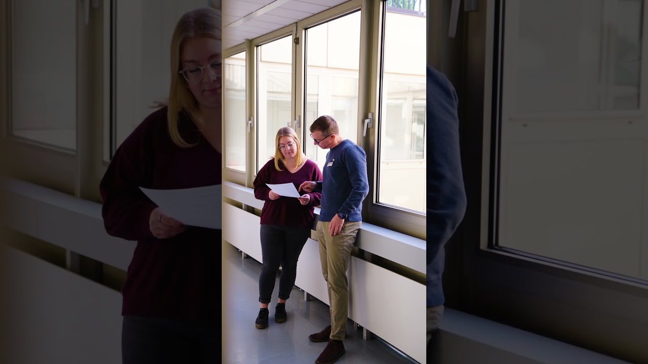 Dieser Alternativtext wurde KI-generiert: Mehrere Personen stehen in einem Flur vor Fenstern. Zwei Personen betrachten Papier in der Hand, während eine dritte Person im Hintergrund steht.