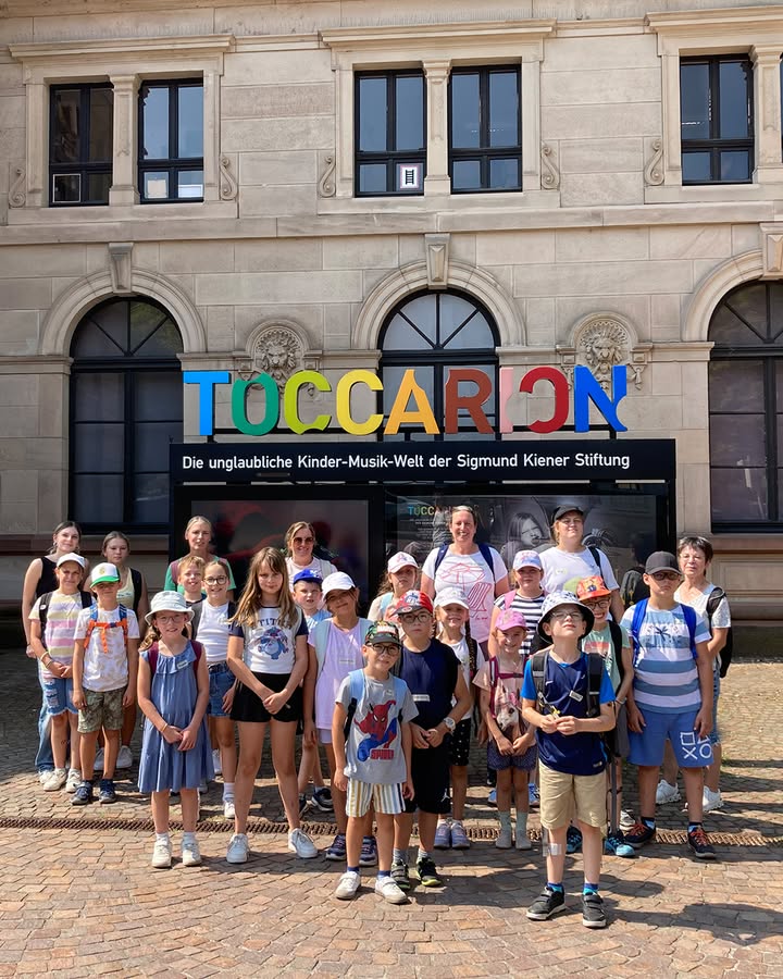Eine große Gruppe von Kindern und einigen Erwachsenen posiert vor einem Steinbauwerk mit dem bunten Schriftzug „TOCCARION“. Darunter ist der Text „Die unglaubliche Kinder-Musik-Welt der Sigmund Kiener Stiftung“ zu lesen. Die Personen blicken in die Kamera. (KI-Alternativtext)
