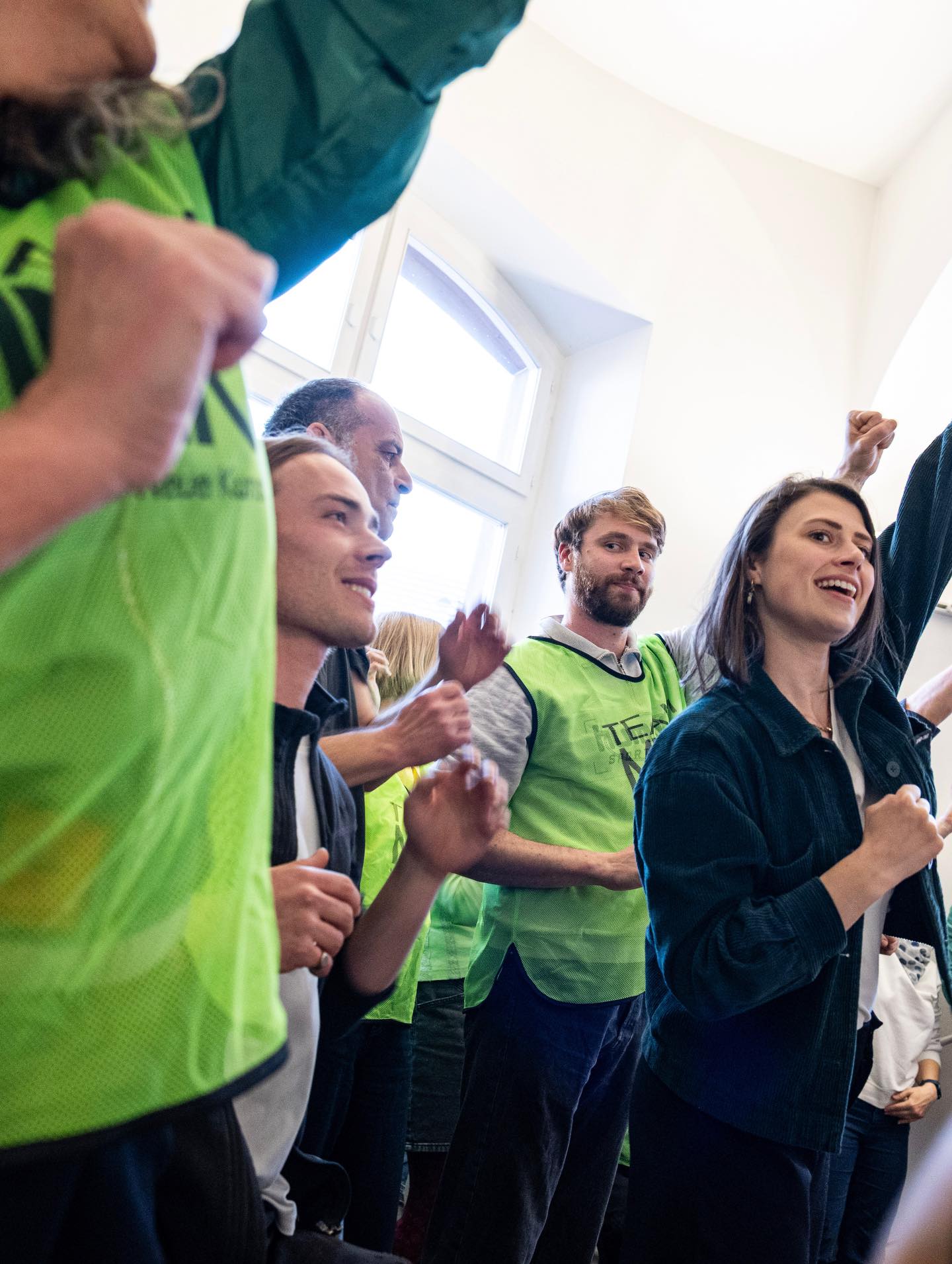 Dieser Alternativtext wurde KI-generiert: Mehrere Personen stehen in einem Raum und haben die Arme erhoben oder klatschen. Sie tragen helle Westen und freuen sich, möglicherweise bei einer Veranstaltung.
