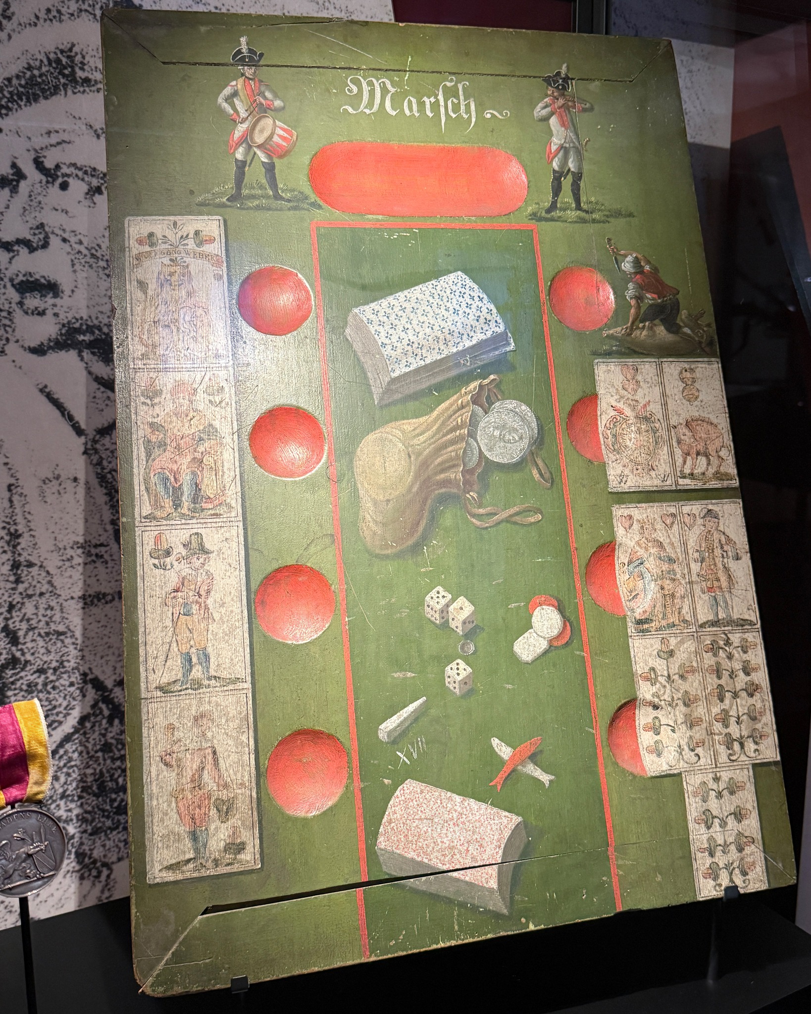 Ein grünes, altes Spielbrett mit der Aufschrift "Marsch" oben in der Mitte. Zwei Personen in Uniform spielen oben Instrumente, während im Zentrum rote Kreise einen Pfad markieren, der von Spielkarten, Würfeln, Münzen und Büchern flankiert wird. (KI-Alternativtext)