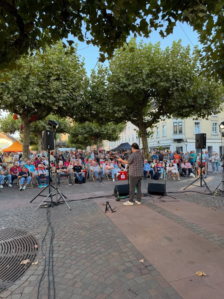 Eine Person steht mit dem Rücken zur Kamera auf einer kleinen Plattform und spielt Gitarne vor einem großen, sitzenden Publikum. Das Publikum ist im Freien auf Stühlen unter Bäumen auf einem gepflasterten Platz versammelt. Lautsprecher und Kabel liegen auf dem Boden. (KI-Alternativtext)