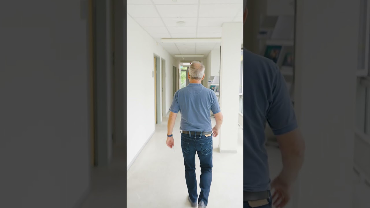 Dieser Alternativtext wurde KI-generiert: Ein Flur mit mehreren Türen erstreckt sich in die Ferne. Im Vordergrund ist der Rücken einer Person zu sehen, die den Flur entlang geht.