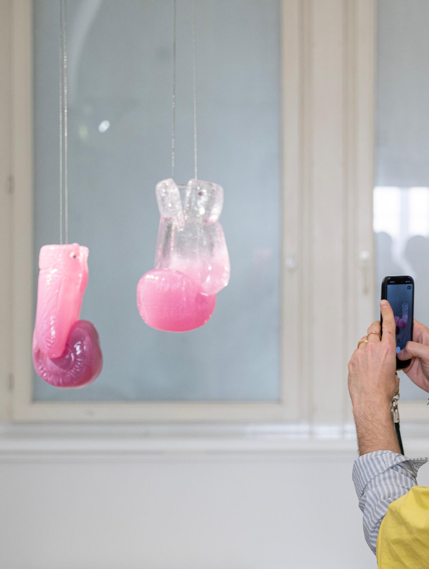 Dieser Alternativtext wurde KI-generiert: Ein oder mehrere rosafarbene Objekte hängen an feinen Fäden vor einem Fenster. Eine Hand hält ein Smartphone, um die Objekte zu fotografieren.