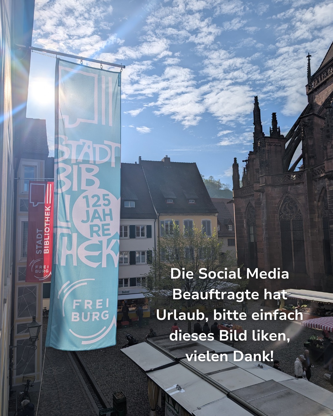 Ein großes hellblaues Banner mit der Aufschrift „STADT BIBLIOTHEK 125 JAHRE FREIBURG“ hängt neben einem roten Banner an einem Gebäude. Im Hintergrund sind historische Häuser und eine gotische Kirche über einem Platz mit Marktständen zu sehen. Ein weißer Text-Overlay lautet: „Die Social Media Beauftragte hat Urlaub, bitte einfach dieses Bild liken, vielen Dank!“ (KI-Alternativtext)
