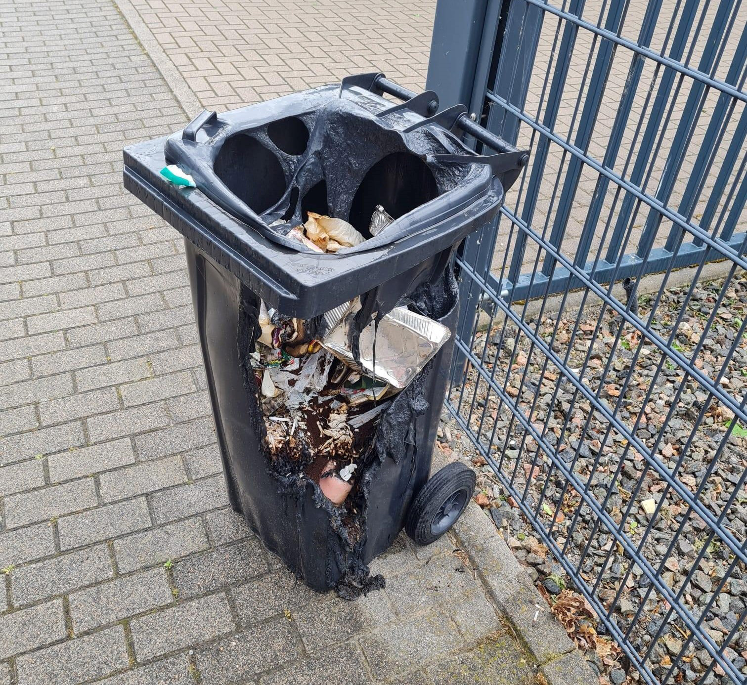 Schwarzer Mülleimer steht auf gepflastertem Boden neben einem Metallzaun. Die Vorderseite ist stark verbrannt und aufgerissen, sodass der Müllinhalt, darunter ein silbernes Blech, sichtbar ist. (KI-Alternativtext)