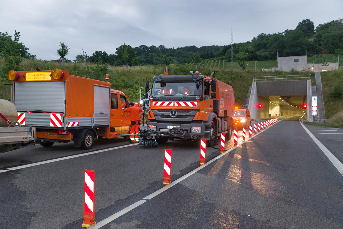Dieser Alternativtext wurde KI-generiert: Ein oder mehrere orangefarbene Arbeitsfahrzeuge stehen auf einer Straße neben einer Baustelle. Sicherheitsvorkehrungen wie Leitkegel und Absperrungen markieren den Arbeitsbereich.