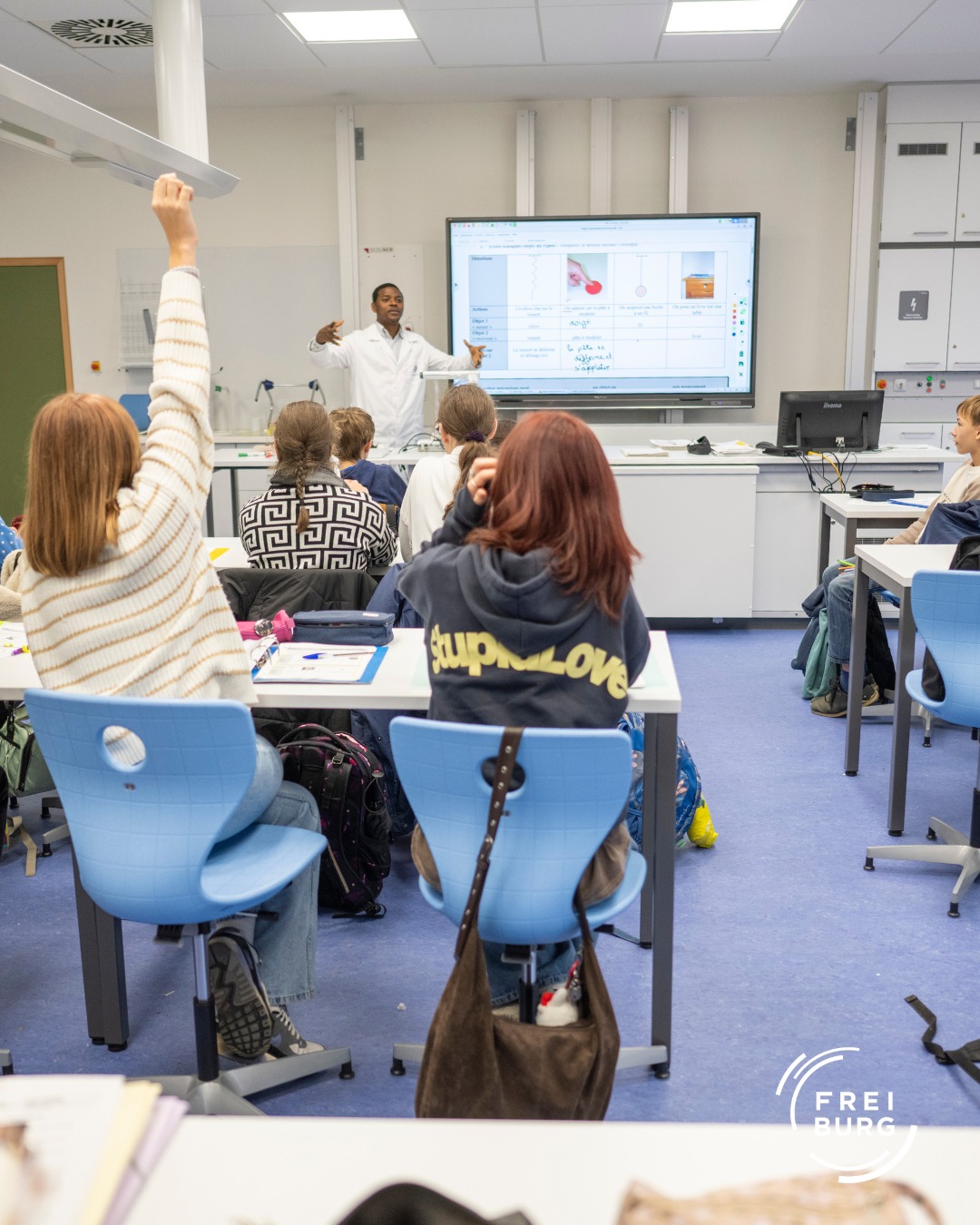 Dieser Alternativtext wurde KI-generiert: Eine Gruppe von Personen sitzt in einem hellen Raum mit Tischen und Stühlen. Eine Person steht vorne und zeigt auf eine Präsentation an der Wand.