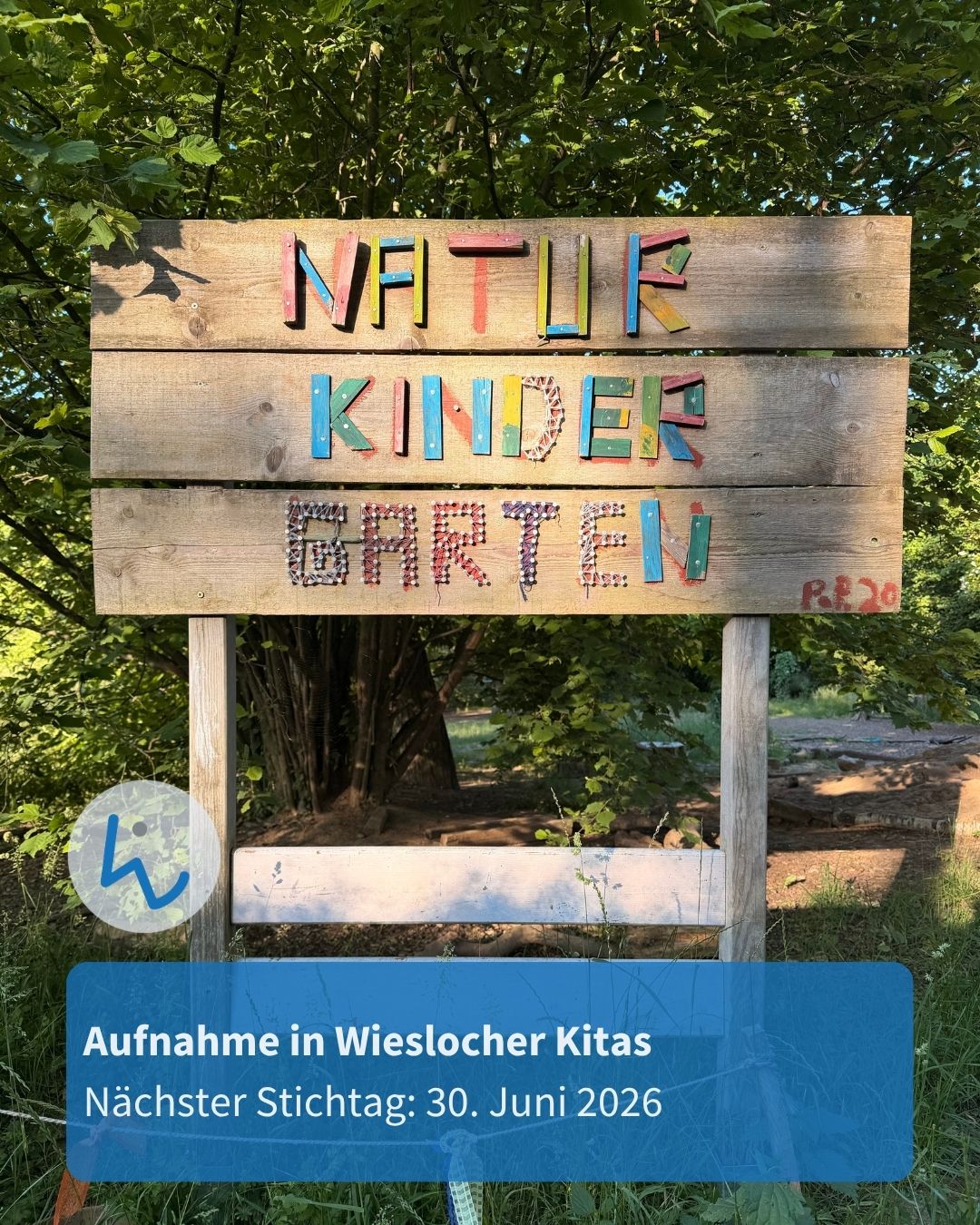 Holzschild mit bunten Buchstaben „NATUR KINDER GARTEN“ vor grüner Vegetation, darunter blauer Balken mit Text: „Aufnahme in Wieslocher Kitas, Nächster Stichtag: 30. Juni 2026“. (KI-Alternativtext)