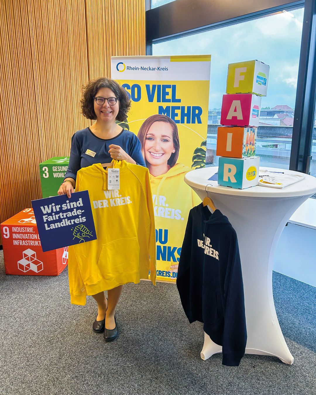 Dieser Alternativtext wurde KI-generiert: Eine Person steht vor einem Infostand mit Bannern und Informationsmaterialien. Im Hintergrund ist ein Gebäude und Tageslicht zu sehen.